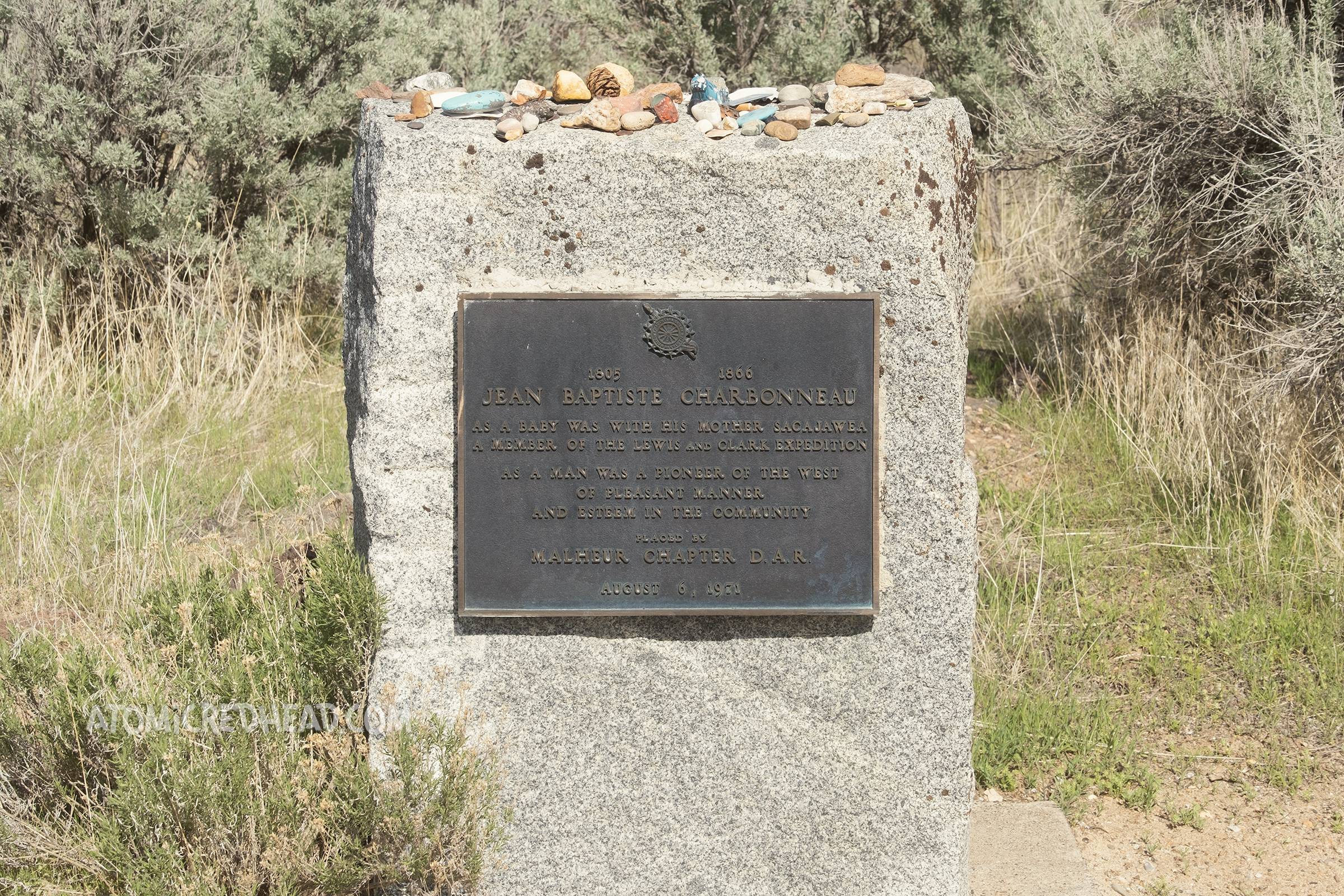 A stone grave marker with a metal plaque reads "1805-1866 Jean Baptiste Charbonneau As a baby was with his mother Sacajawea as a member of the Lewis and Clark Expedition. As a man was a pioneer of the west of pleasant manner and esteem in the community. Placed by Malheau Chapter D.A.R. August 6, 1971.
