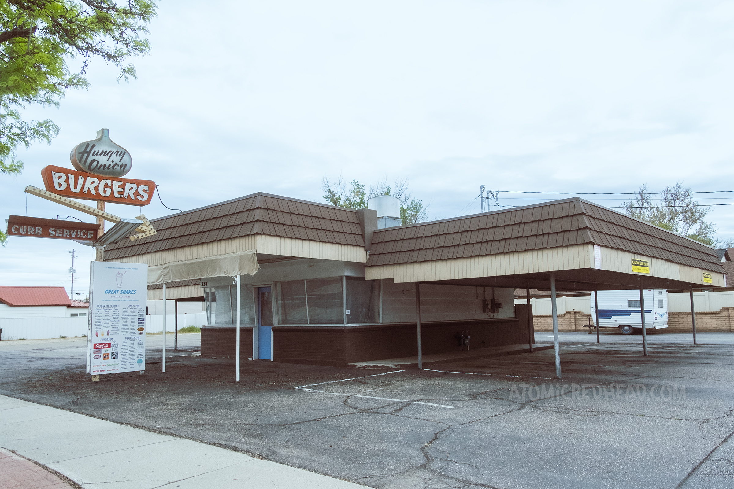 Overall view of a single story drive-in restaurant with a covered area for cars on the right and a walk-up window on the left. A features an onion shape and reads "Hungry Onion" inside, below "Burgers" in neon, and an arrow pointing toward the building.