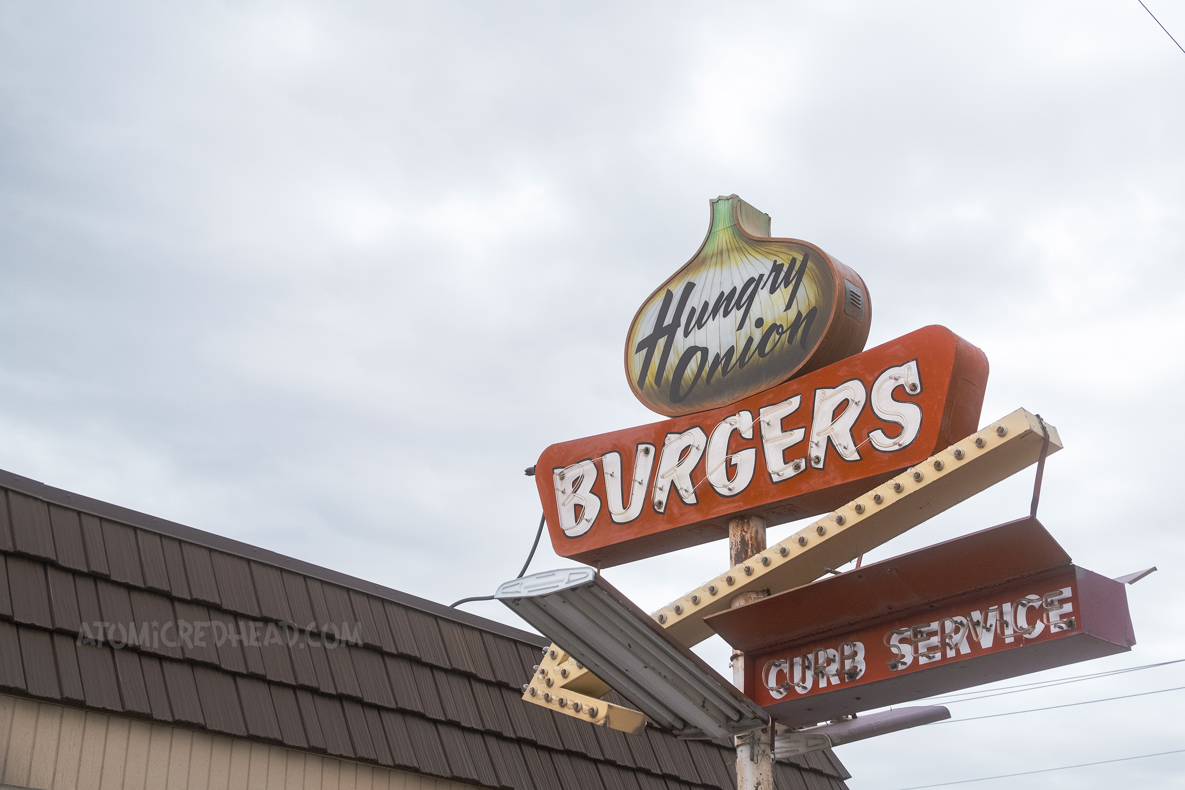 Close-up view of the Hungry Onion sign, upper portion is backlit plastic in the shape of an onion, and reads "Hungry Onion" below is a red and white sign reading "Burgers" in neon, and a yellow arrow below that and below that small neon reads "Curb service"