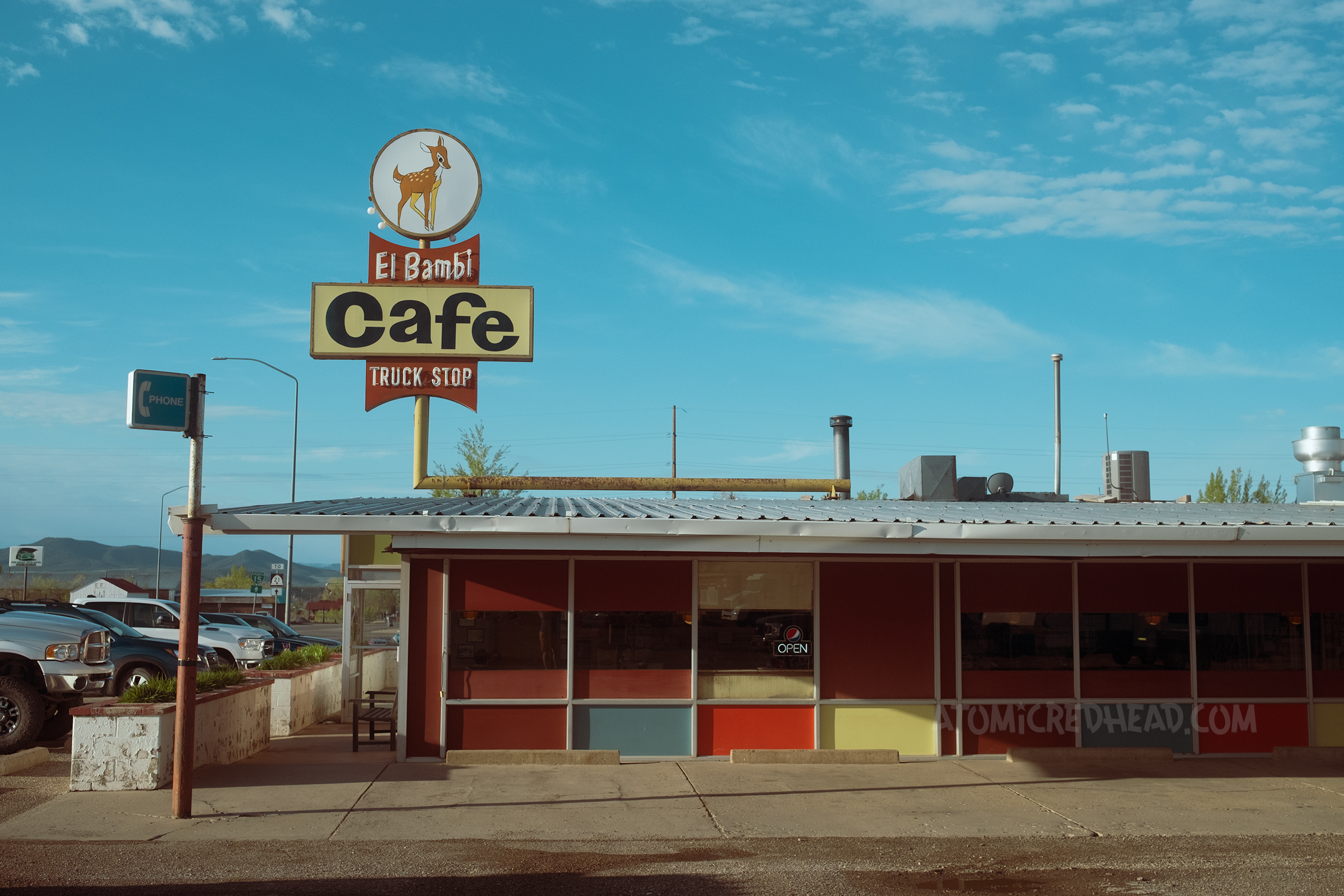 Overall view of the El Bambi, which has large windows and painted in various colors. Atop is a combination backlit plastic and neon sign reading "El Bambi Cafe Truck Stop" with an image of a deer at the top.