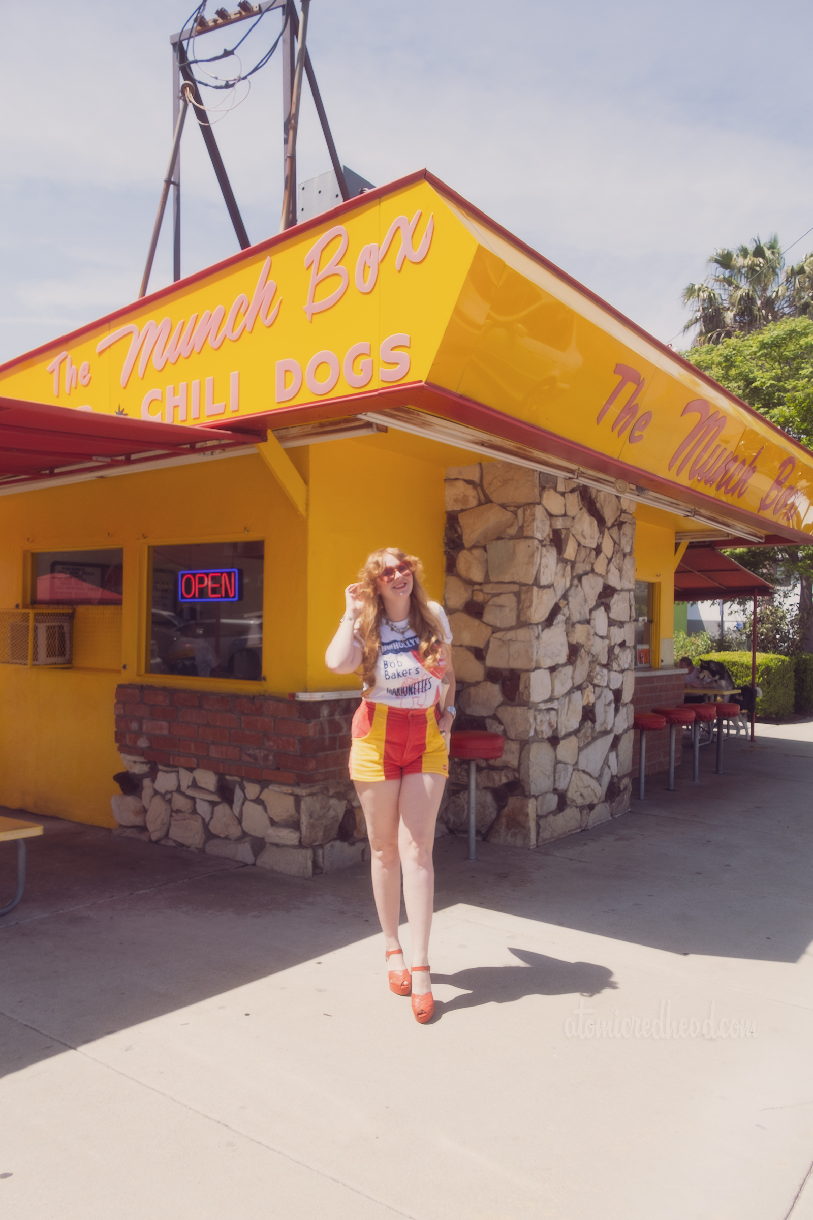 Myself standing outside of the bright yellow and red Munch Box building, wearing a white shirt with a clown marionette on it and text reading "From Hollywood Bob Baker's Marionettes" and red shorts with yellow pockets.
