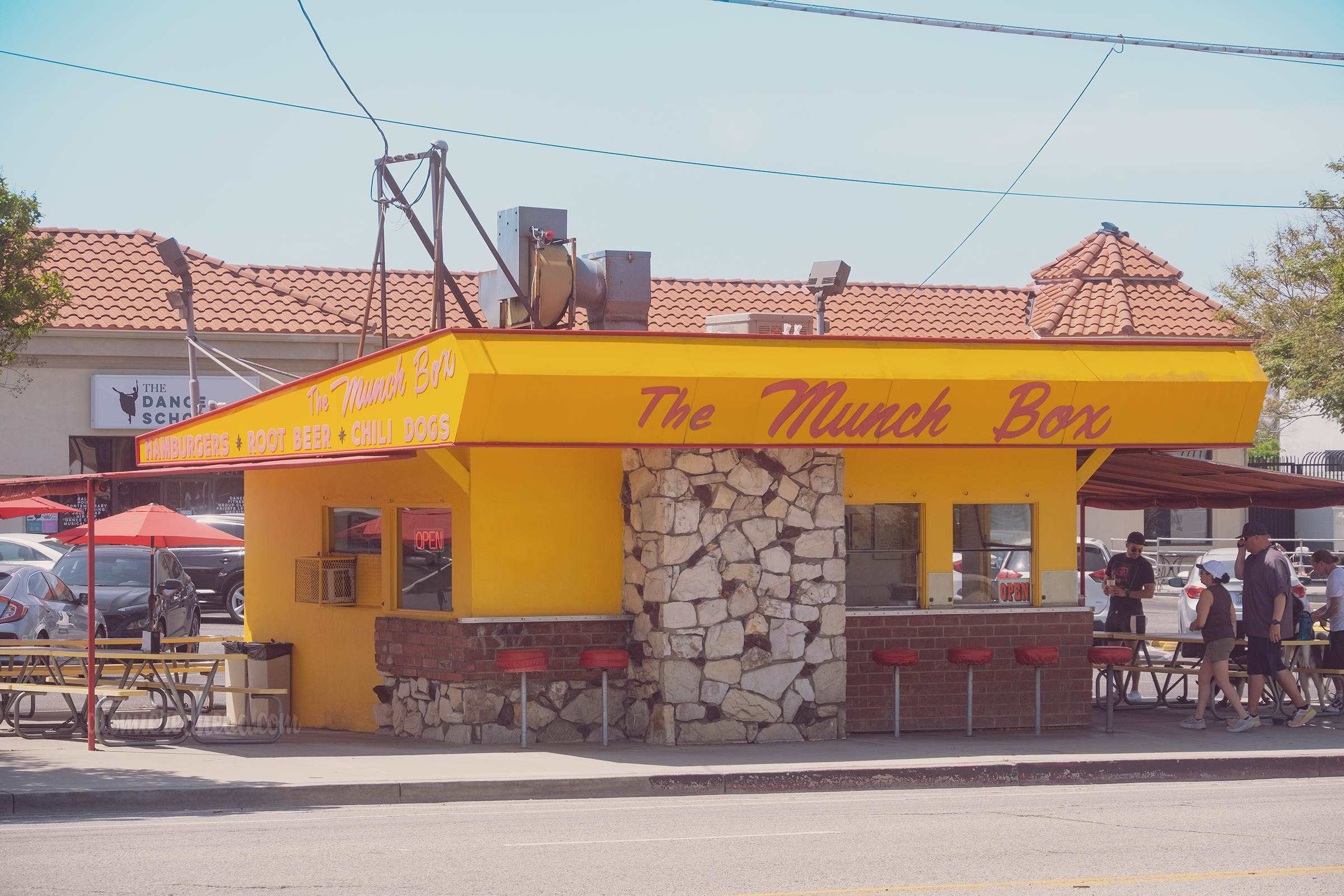 Overall view of the Munch Box, which is a small yellow building with an angled Googie roof, rock wall portion in front, and lower area made up of red brick and red vinyl bar stools. Red script across the roof reads "The Munch Box"