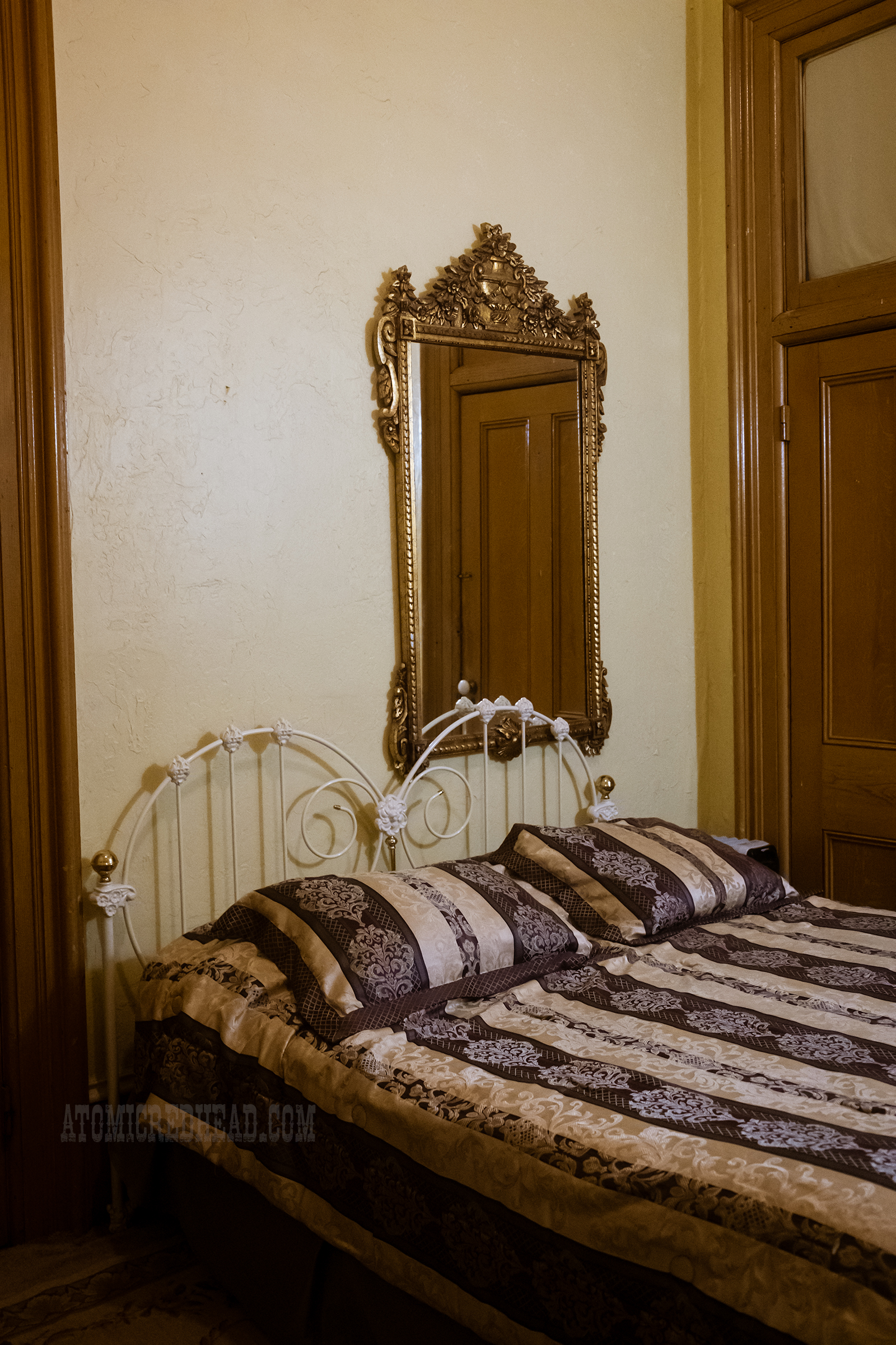 A heart shaped metal headboard rests against a cream wall. The bedspread features a floral stripe pattern of white and maroon. Above the bed hangs a mirror with a gold frame.