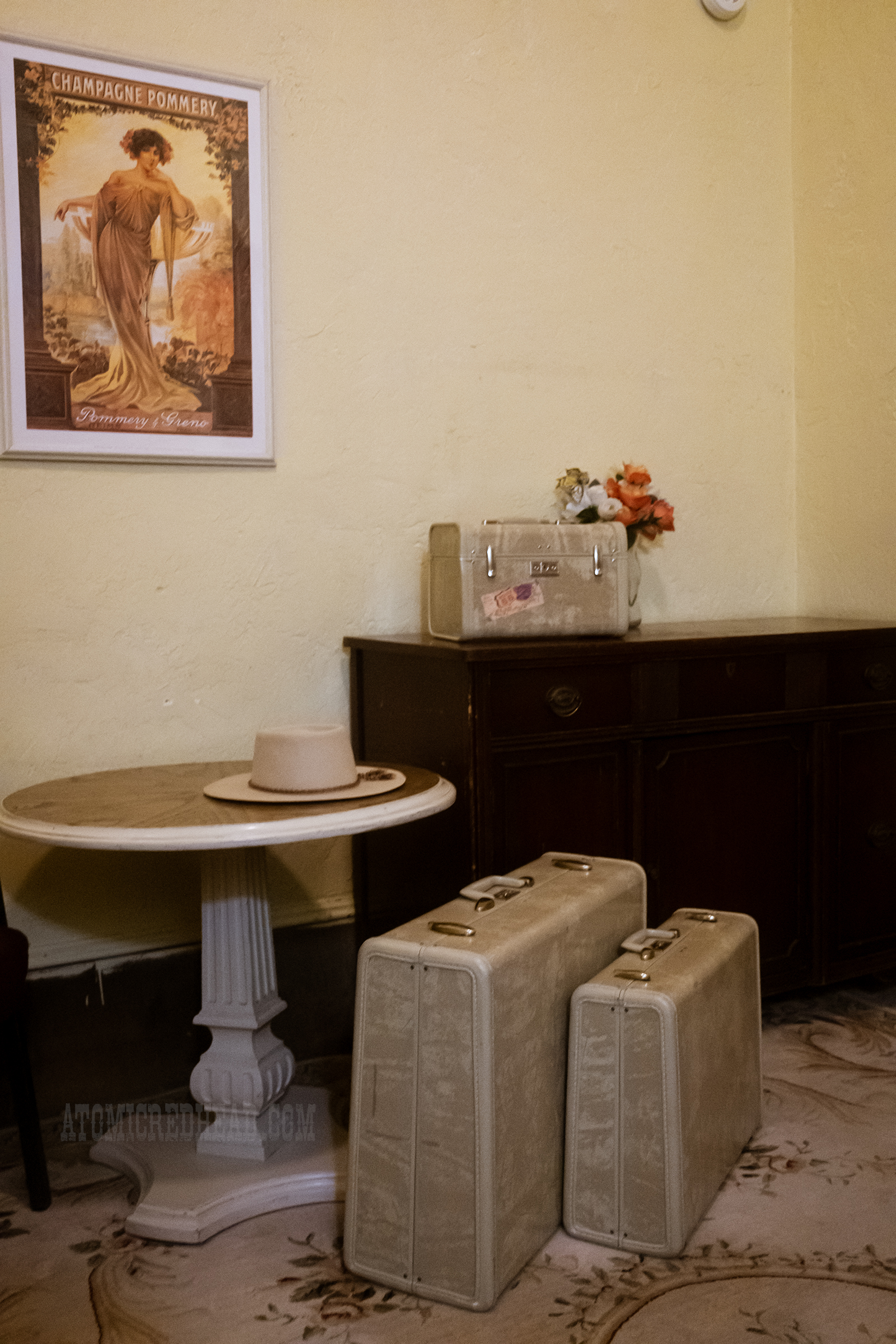 A small dark wood dresser sits against a white wall with a small circular table next to it. Atop the dresser is a cream train case, and on the floor two matching suitcases.