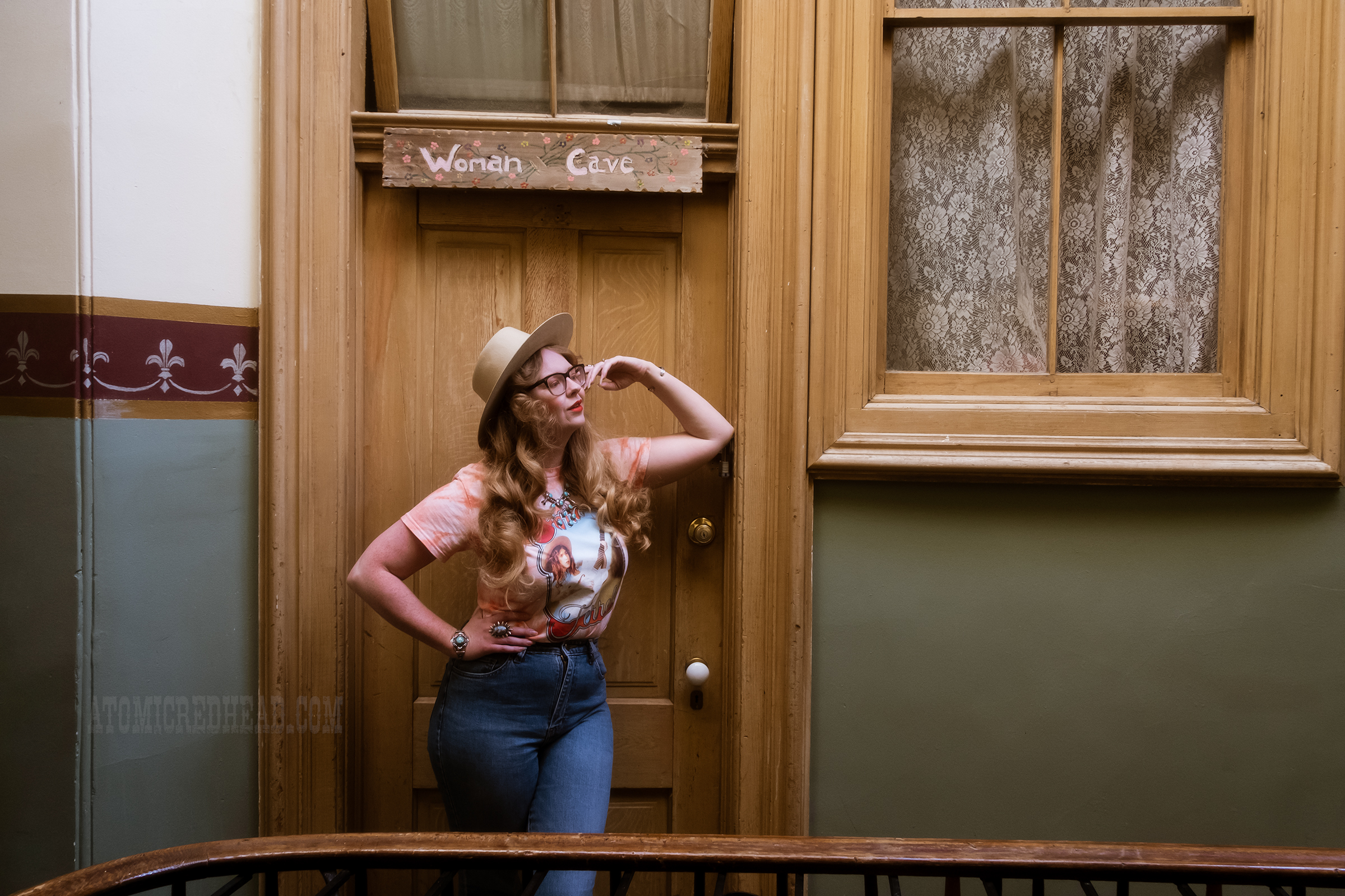 Myself leaning against a doorway with a sign that reads "Woman Cave" above, wearing a cream cowboy hat, peach tie dye shirt featuring musician Sierra Ferrell. 