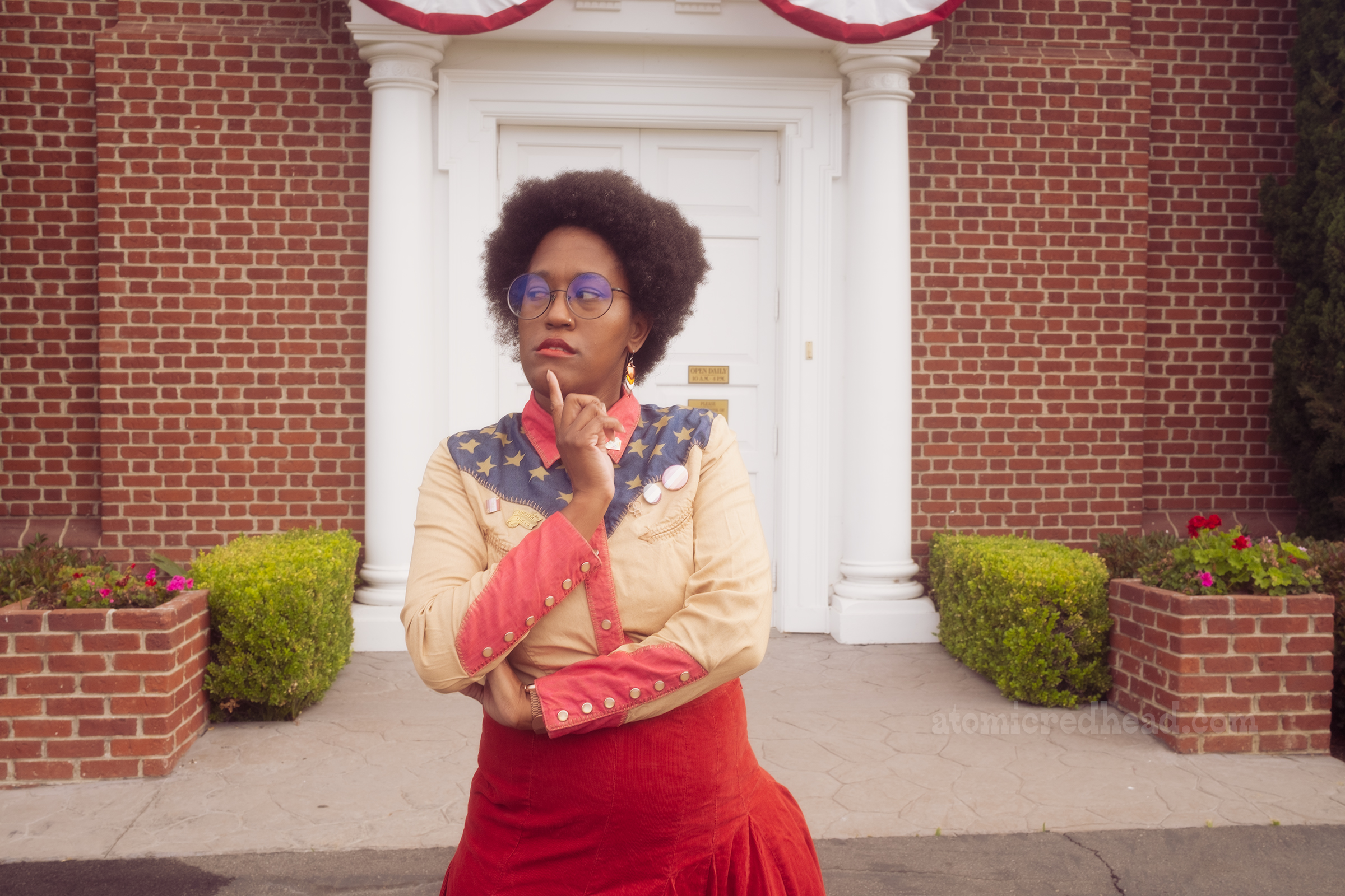 Carla, wearing a western wear shirt with a yoke of blue with white stars, and a pale tan with red cuffs, and a red skirt, stands in front of Independence Hall.