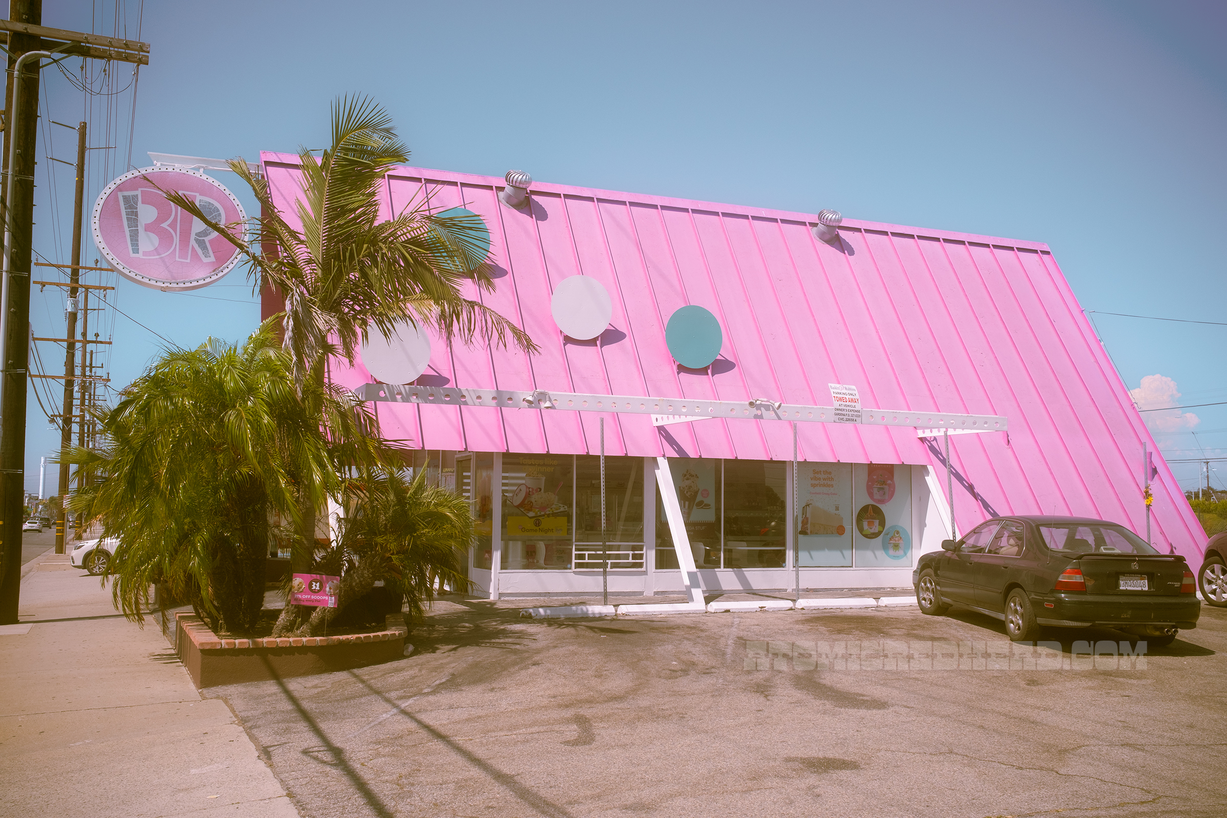 Slightly angled view of the Baskin-Robbins, an A-frame building with a pink roof. White, pink and blue dots stand on the roof, and jutting out from the front is a circular sign that reads "BR" in pink, white, and blue.
