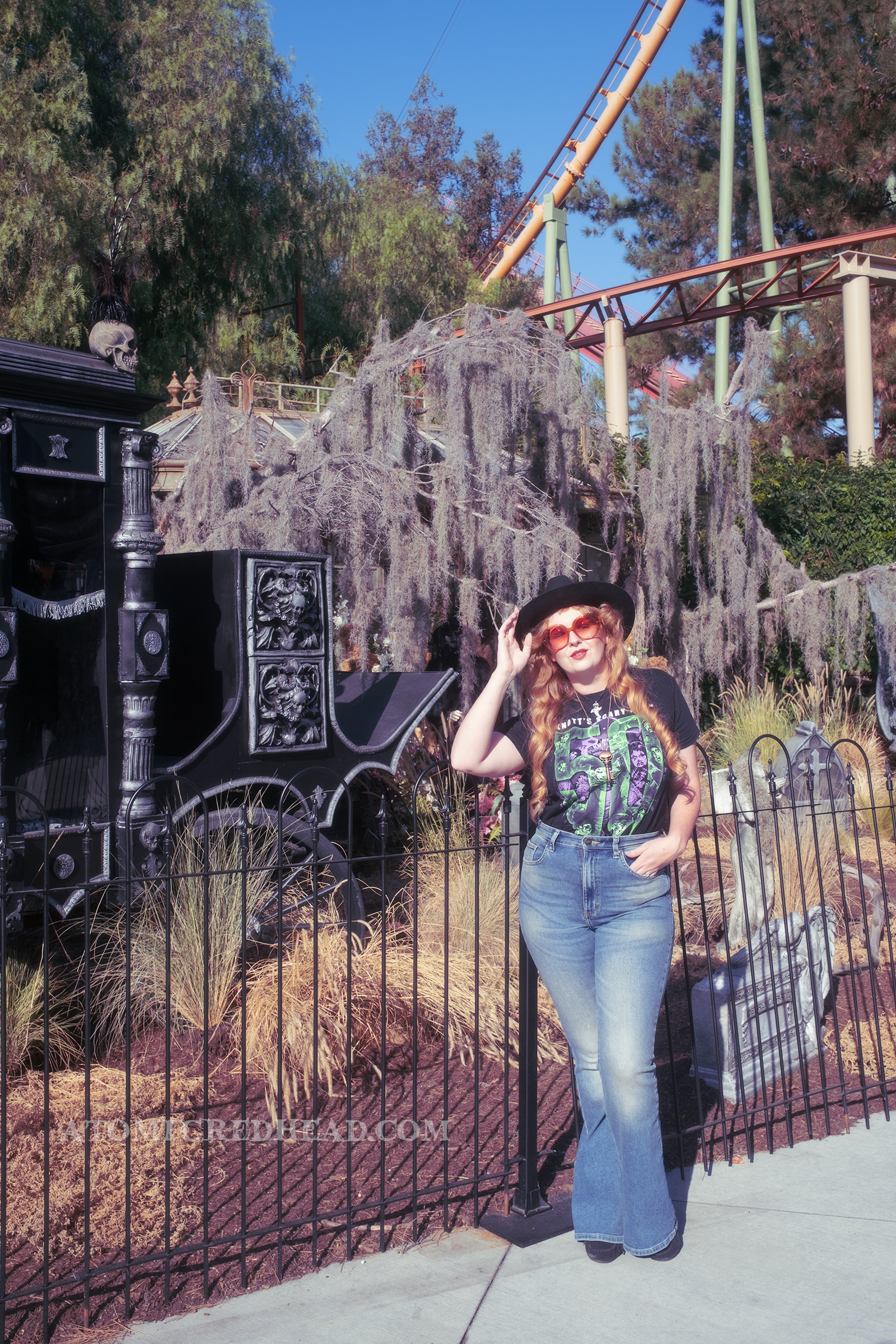 Myself, wearing a black cowboy hat, a black shirt that features various Scary Farm Monsters and reads "50" in big green numbers, and jeans, standing against a fence with a cemetery scene behind.