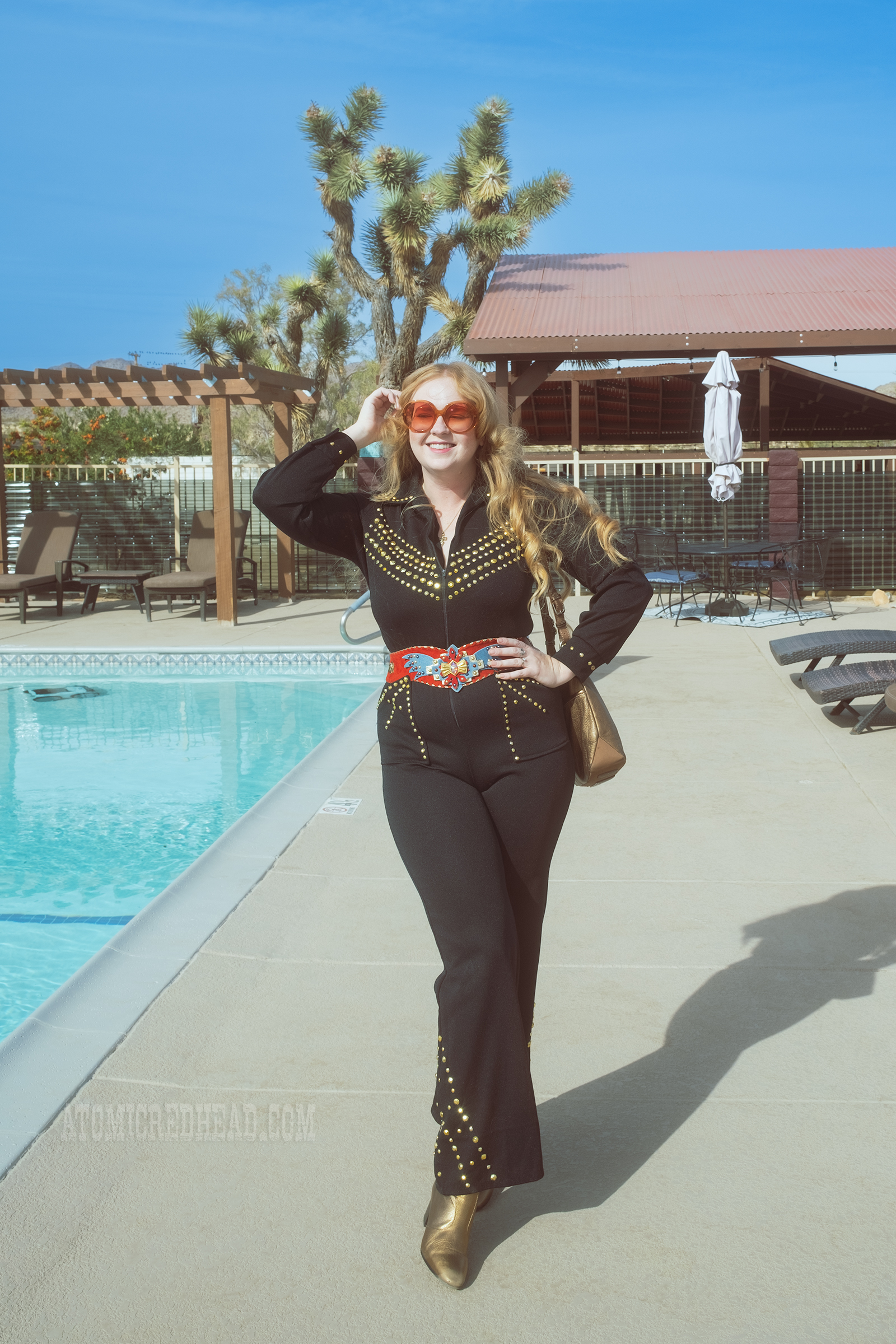 Myself, wearing a black jumpsuit, with gold rhinestones, a red belt with an eagle on it, and gold boots, standing in front of a pool.