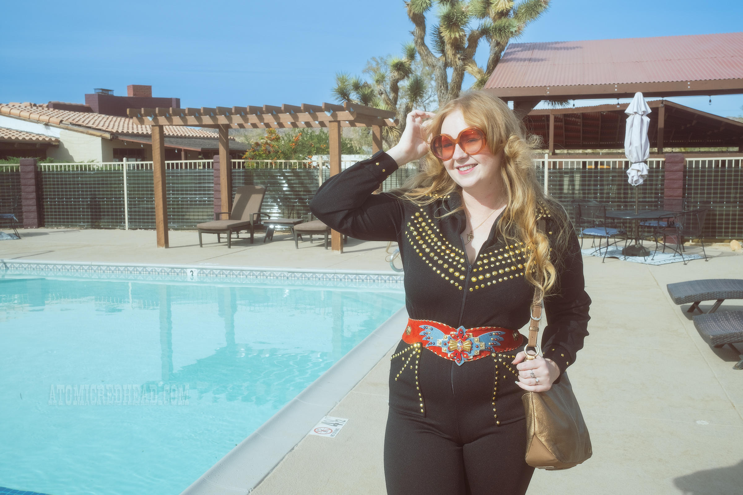 Myself, wearing a black jumpsuit, with gold rhinestones, a red belt with an eagle on it, and gold boots, standing in front of a pool.