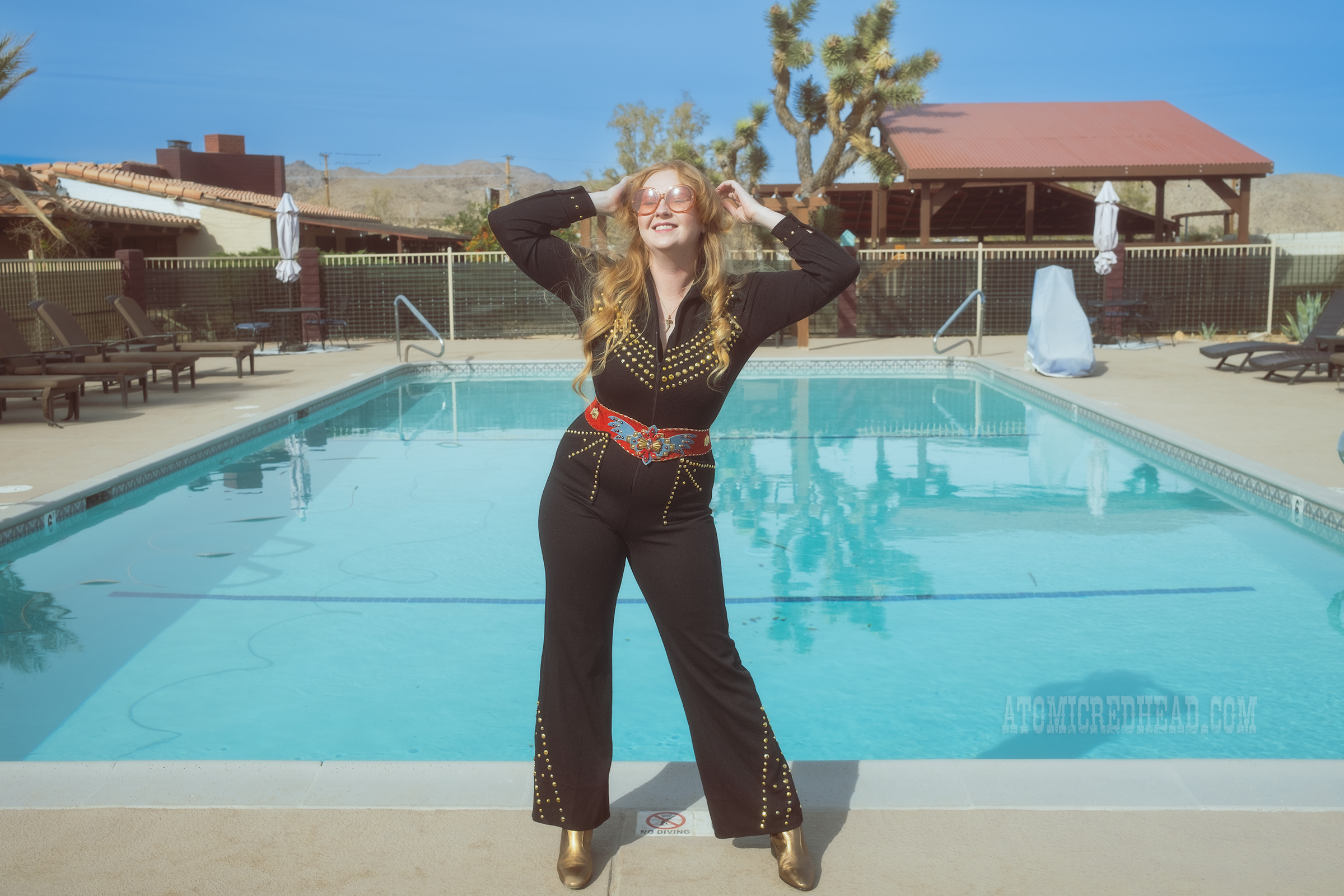 Myself, wearing a black jumpsuit, with gold rhinestones, a red belt with an eagle on it, and gold boots, standing in front of a pool.