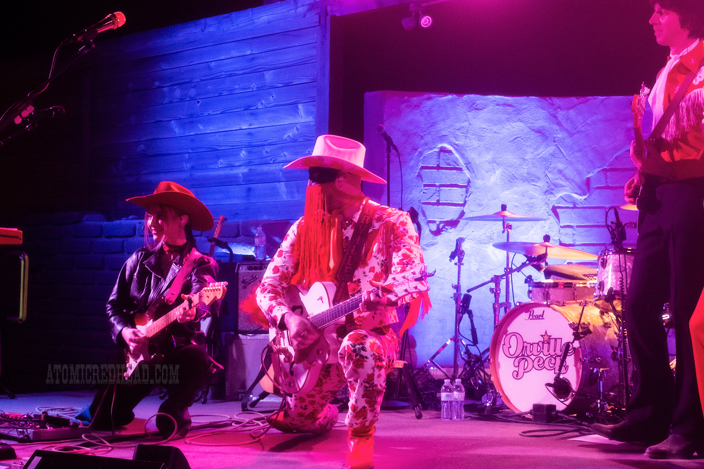 Orville Peck, wearing a white outfit with roses printed all over it, a white cowboy hat, and a black mask with red fringe, performs.