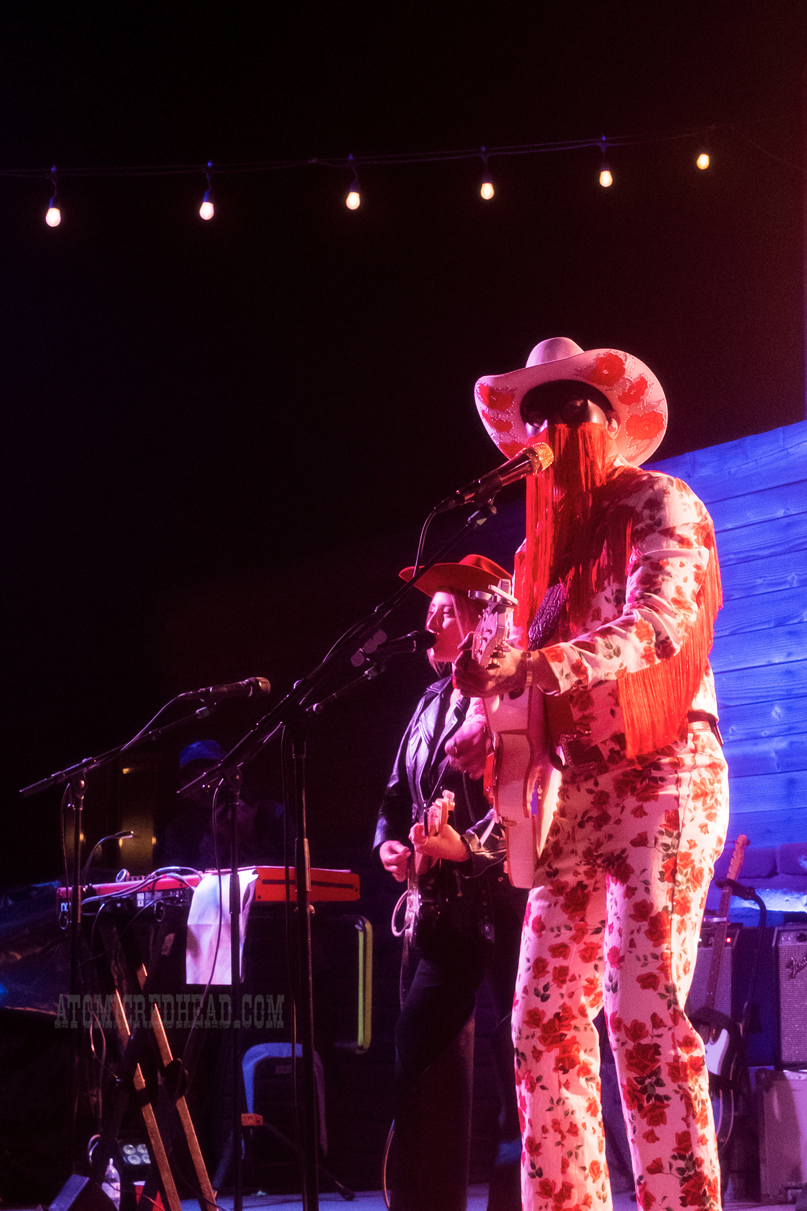 Orville Peck, wearing a white outfit with roses printed all over it, a white cowboy hat, and a black mask with red fringe, performs.