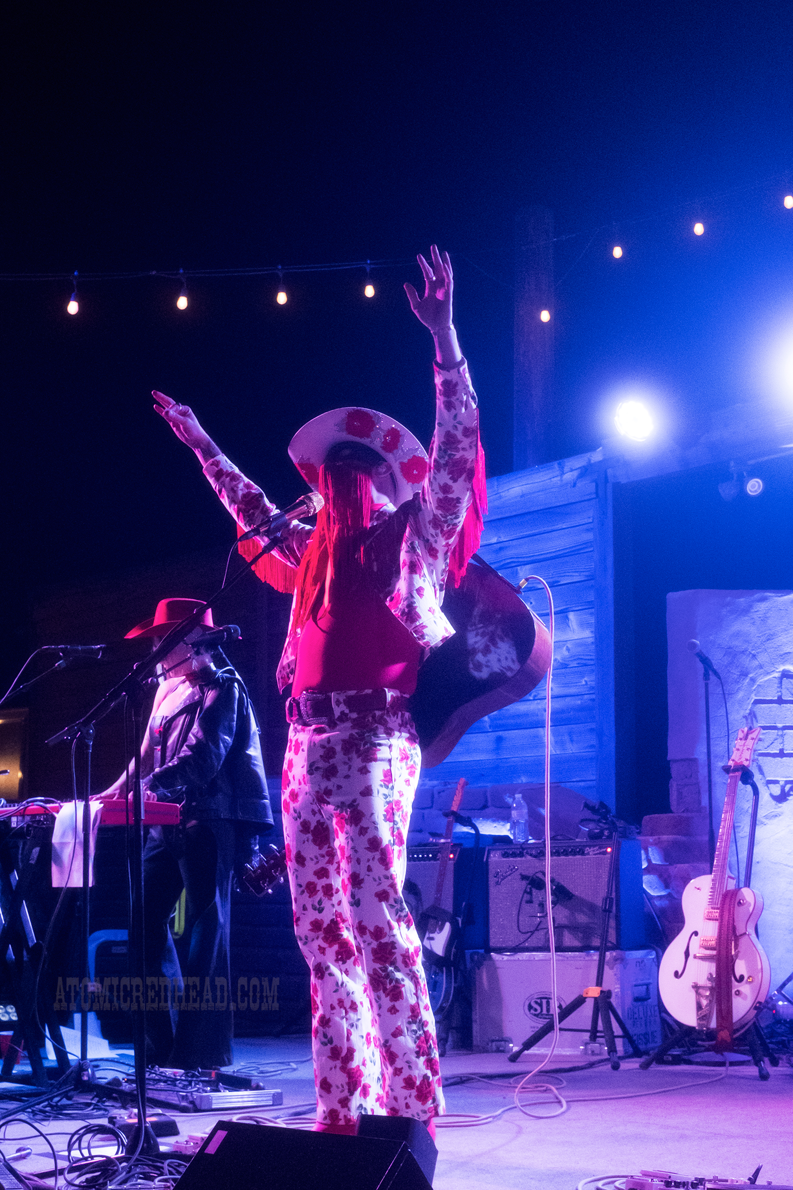 Orville Peck, wearing a white outfit with roses printed all over it, a white cowboy hat, and a black mask with red fringe, performs.