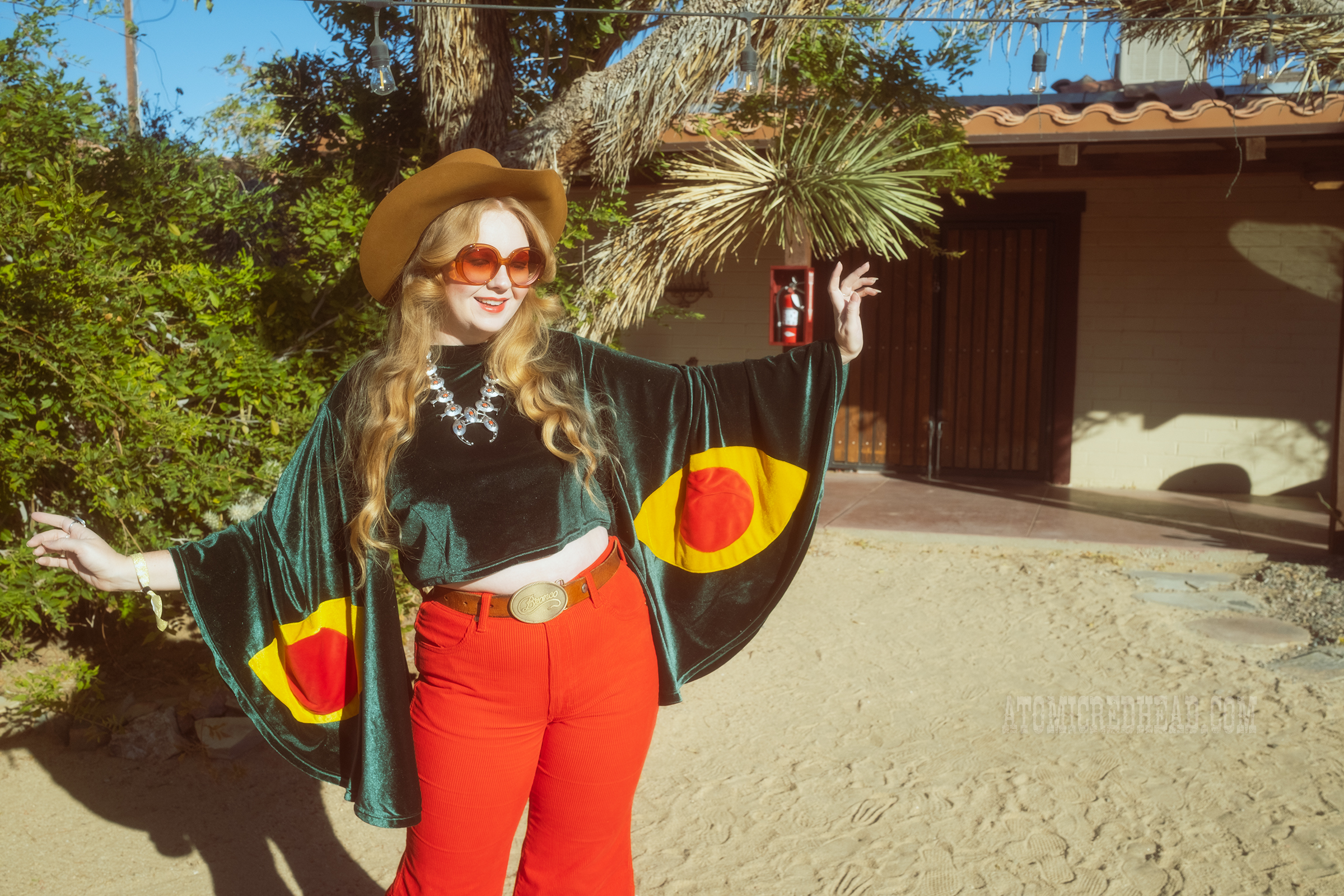 Myself, wearing a light brown cowboy hat, a green velvet top with bell sleeves and red and yellow eye shapes on the sleeves, red corduroy pants, and green shoes.