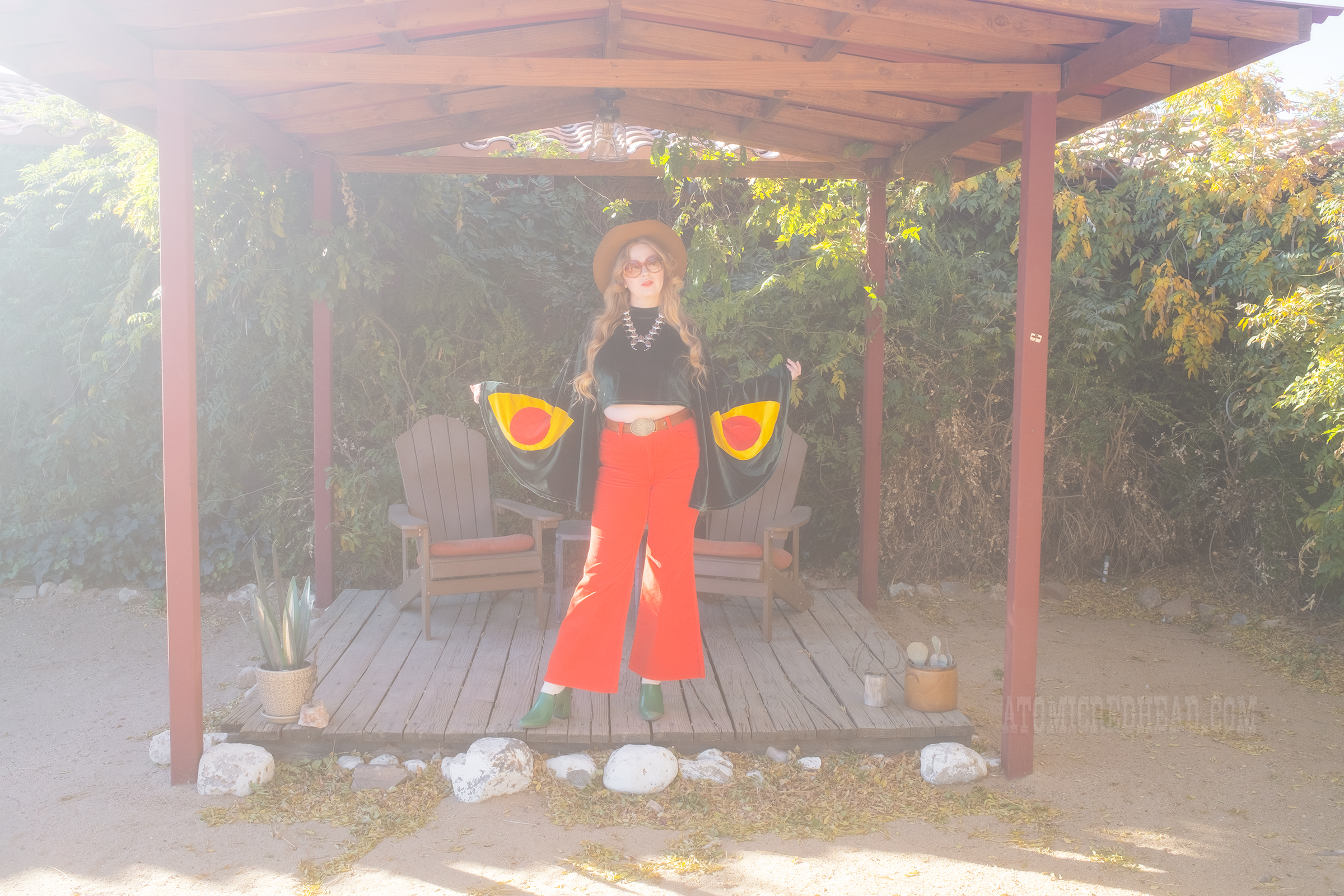 Myself, wearing a light brown cowboy hat, a green velvet top with bell sleeves and red and yellow eye shapes on the sleeves, red corduroy pants, and green shoes.
