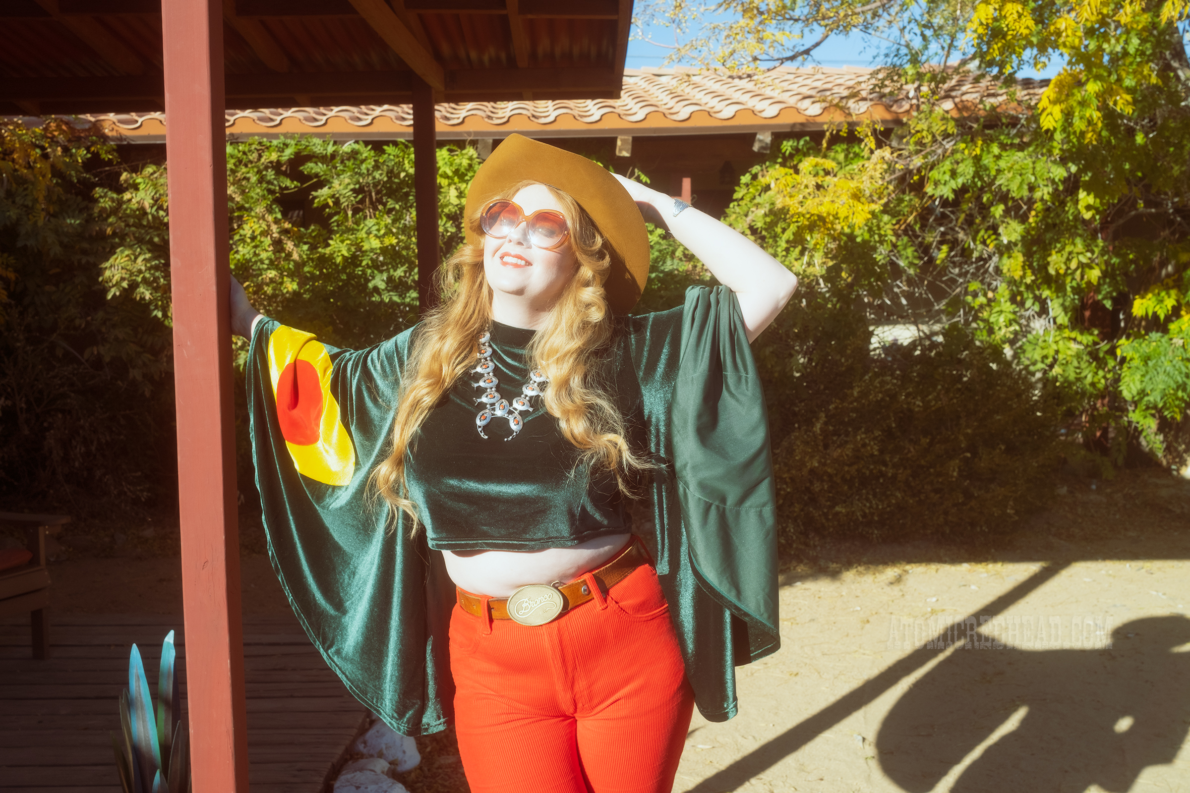 Myself, wearing a light brown cowboy hat, a green velvet top with bell sleeves and red and yellow eye shapes on the sleeves, red corduroy pants, and green shoes.