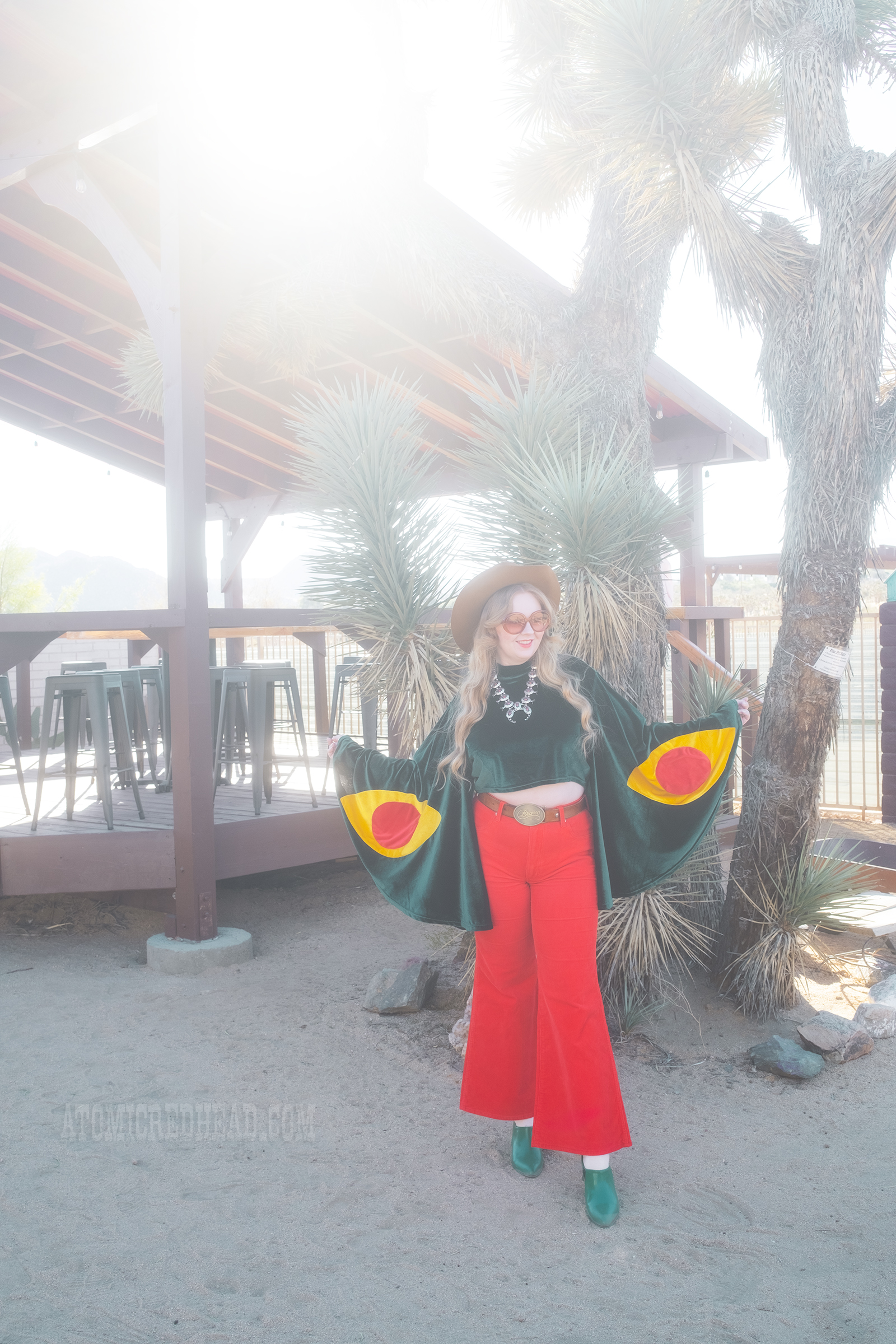 Myself, wearing a light brown cowboy hat, a green velvet top with bell sleeves and red and yellow eye shapes on the sleeves, red corduroy pants, and green shoes.