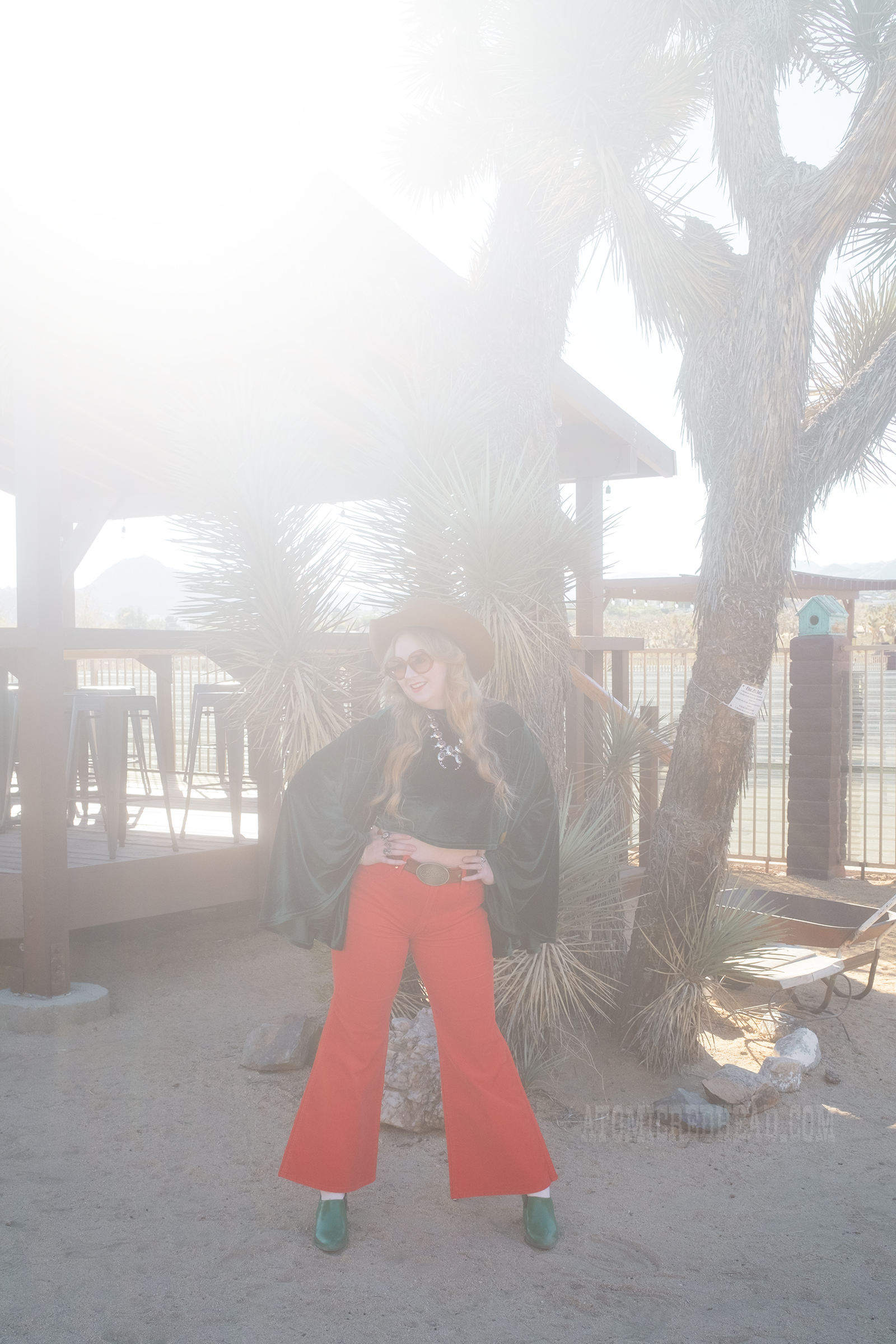 Myself, wearing a light brown cowboy hat, a green velvet top with bell sleeves and red and yellow eye shapes on the sleeves, red corduroy pants, and green shoes.
