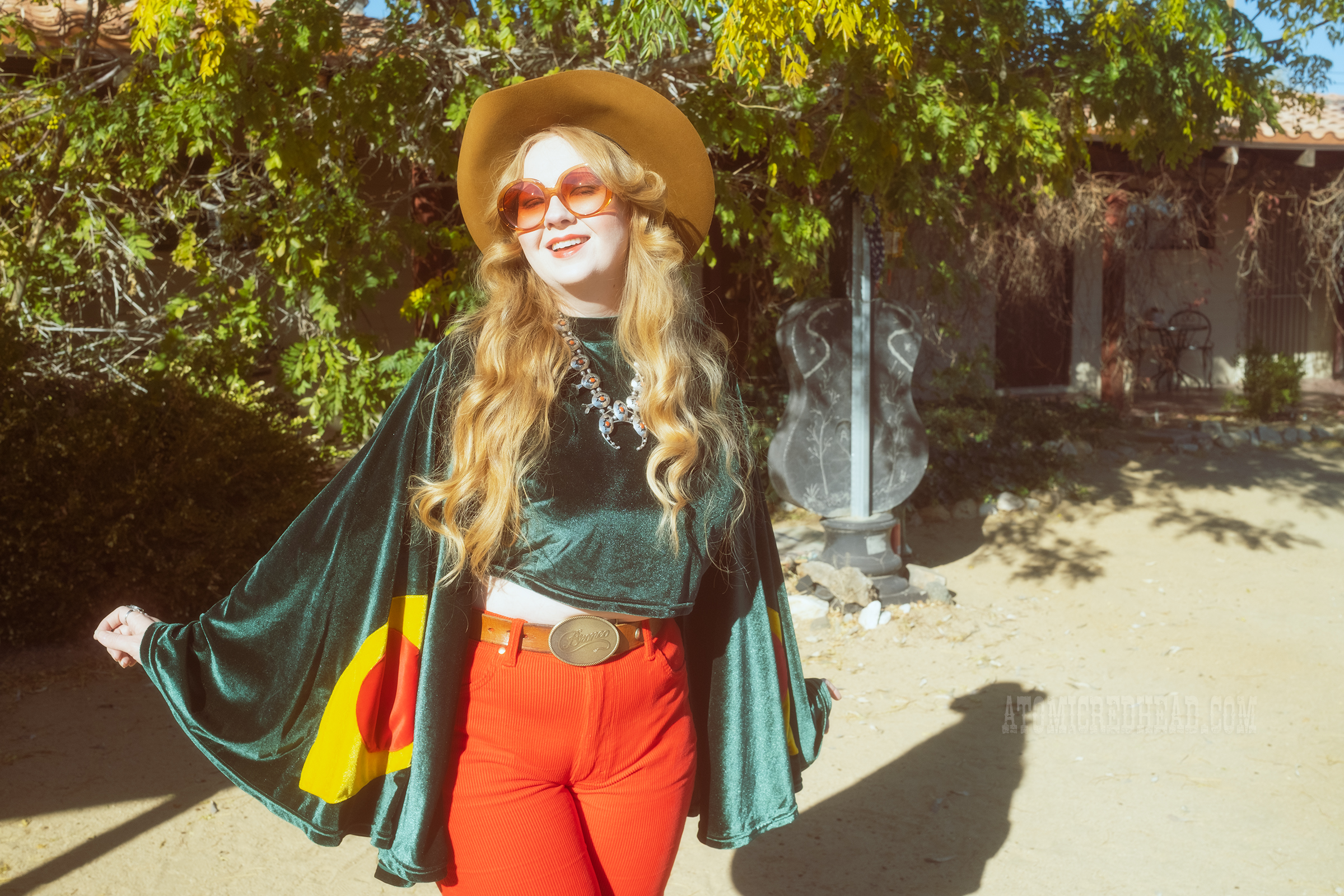 Myself, wearing a light brown cowboy hat, a green velvet top with bell sleeves and red and yellow eye shapes on the sleeves, red corduroy pants, and green shoes.