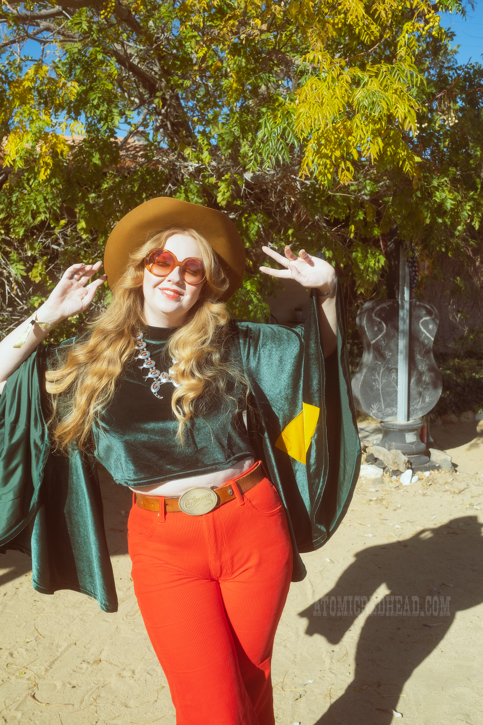 Myself, wearing a light brown cowboy hat, a green velvet top with bell sleeves and red and yellow eye shapes on the sleeves, red corduroy pants, and green shoes.