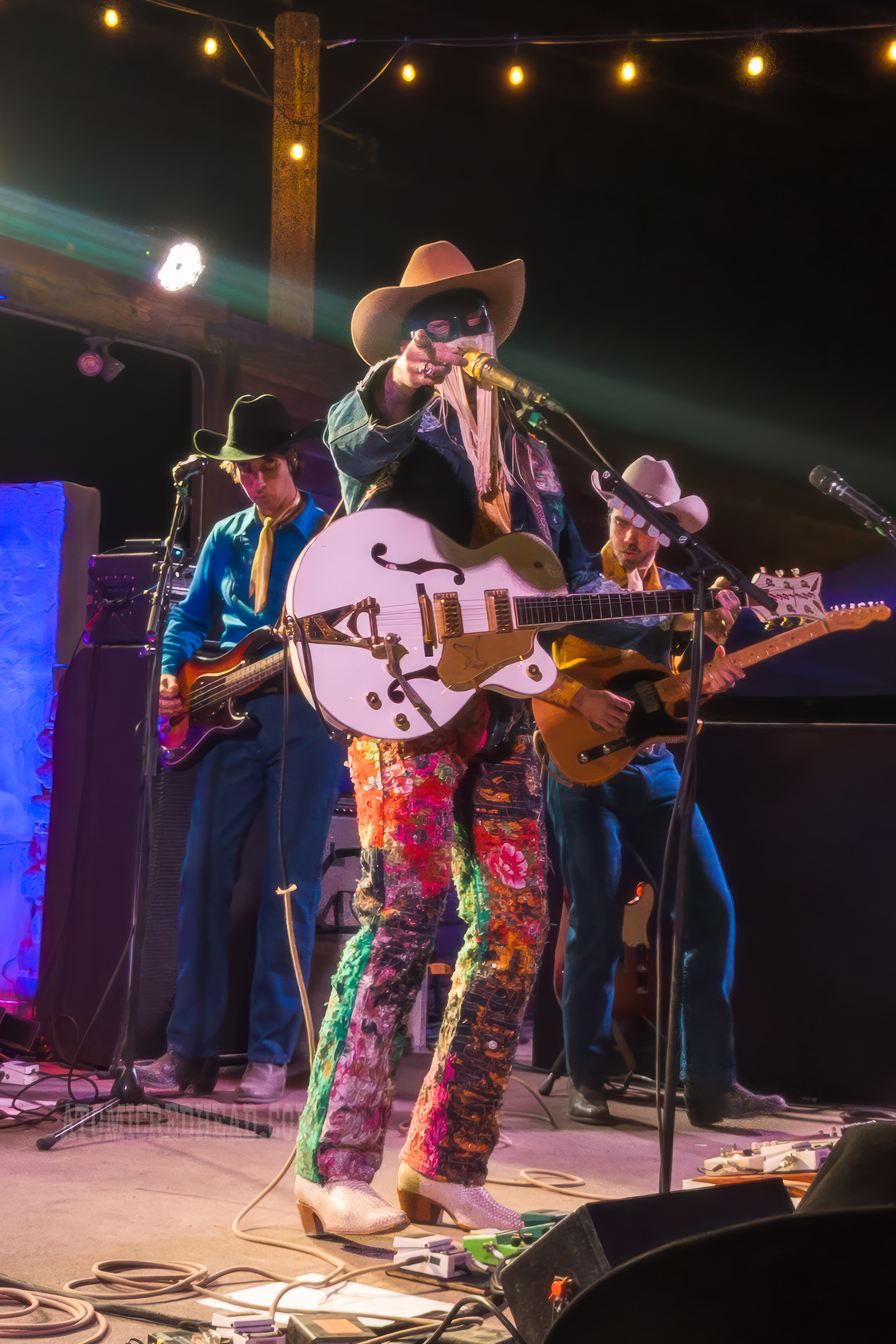 Orville Peck, wearing a jean jacket and jean pants covered in bandanas plays the guitar and points to the camera.