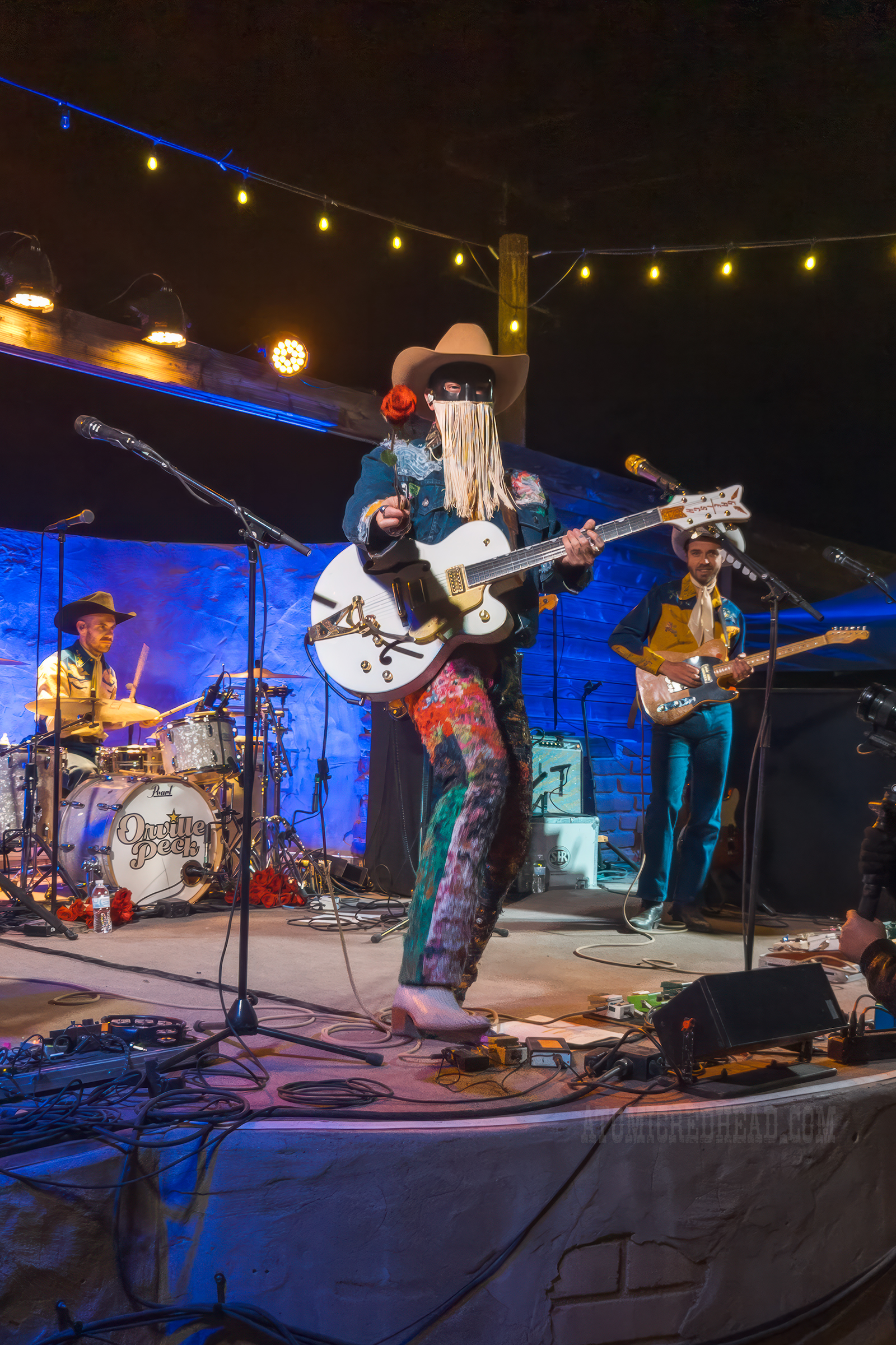Orville Peck, wearing a jean jacket and jean pants covered in bandanas holds out a rose.