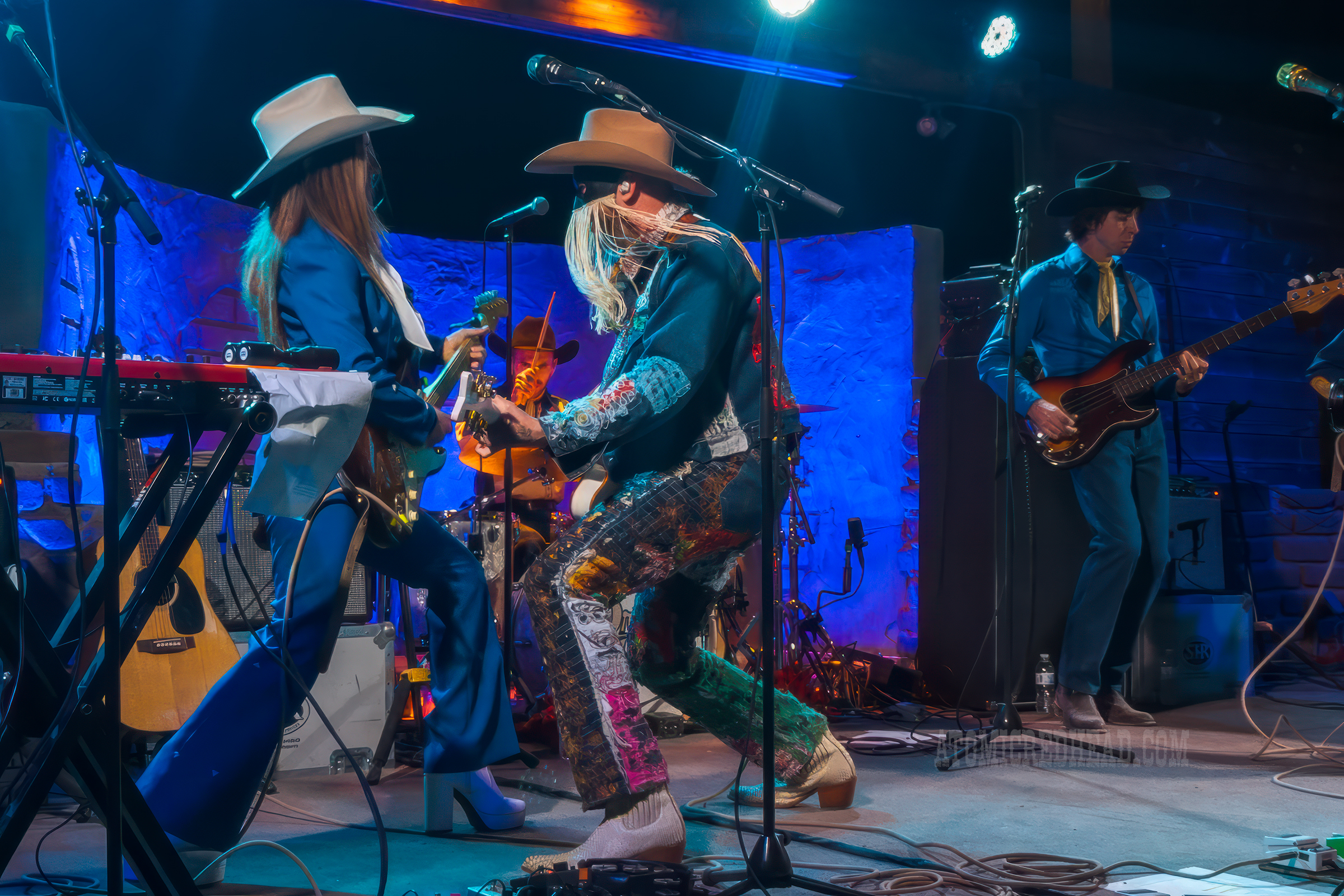 Orville Peck, wearing a jean jacket and jean pants covered in bandanas plays the guitar