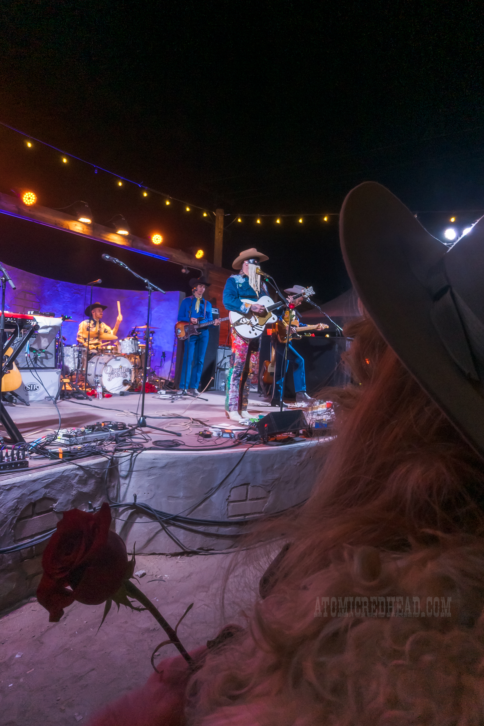 Orville Peck, wearing a jean jacket and jean pants covered in bandanas plays the guitar. In the foreground is me, holding my rose.