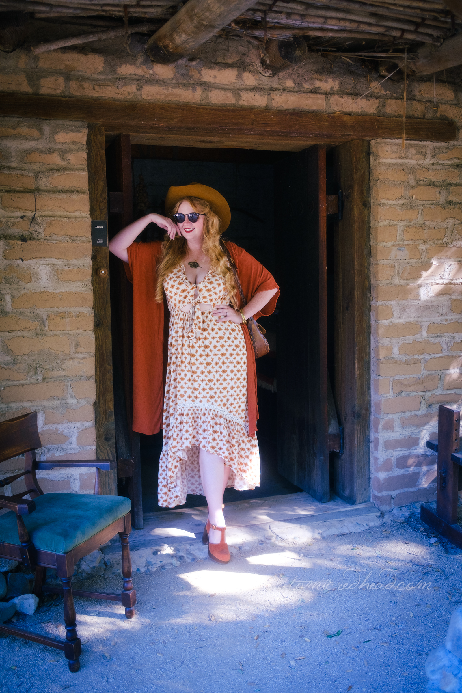 Myself, leaning against the doorway of the adobe wearing a brown cowboy hat, brown wrap, and a long dress of cream and brown.