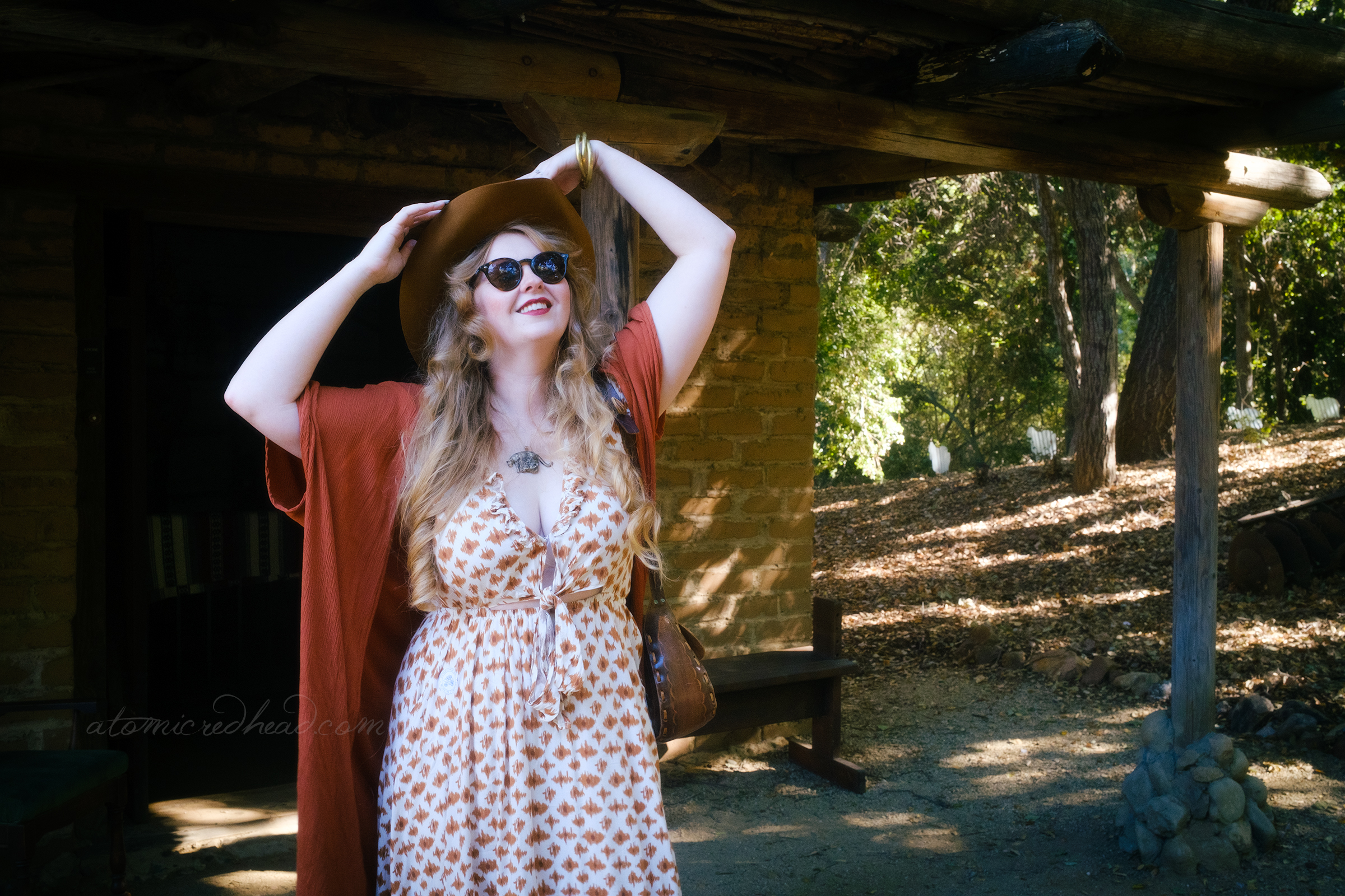 Myself, leaning against the doorway of the adobe wearing a brown cowboy hat, brown wrap, and a long dress of cream and brown.