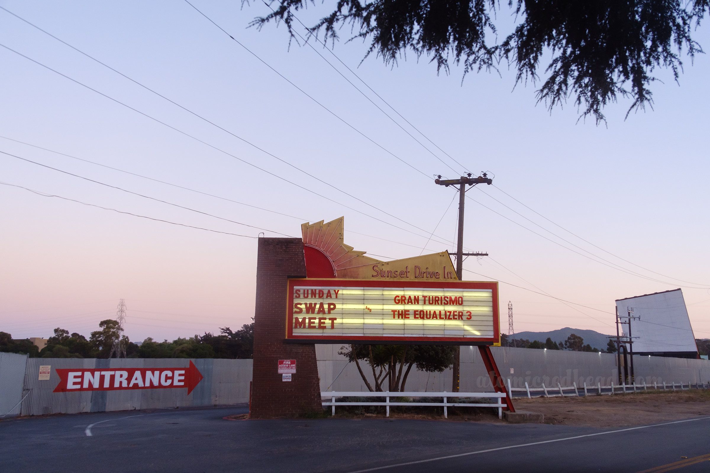Marquee, featuring a neon sun, and neon text reading "Sunset Drive-In" on the marquee it reads "Sunday Swap Meet Gran Turismo and The Equalizer 3" in the distance, painted on a corrugated metal wall in red and white reads "Entrance"