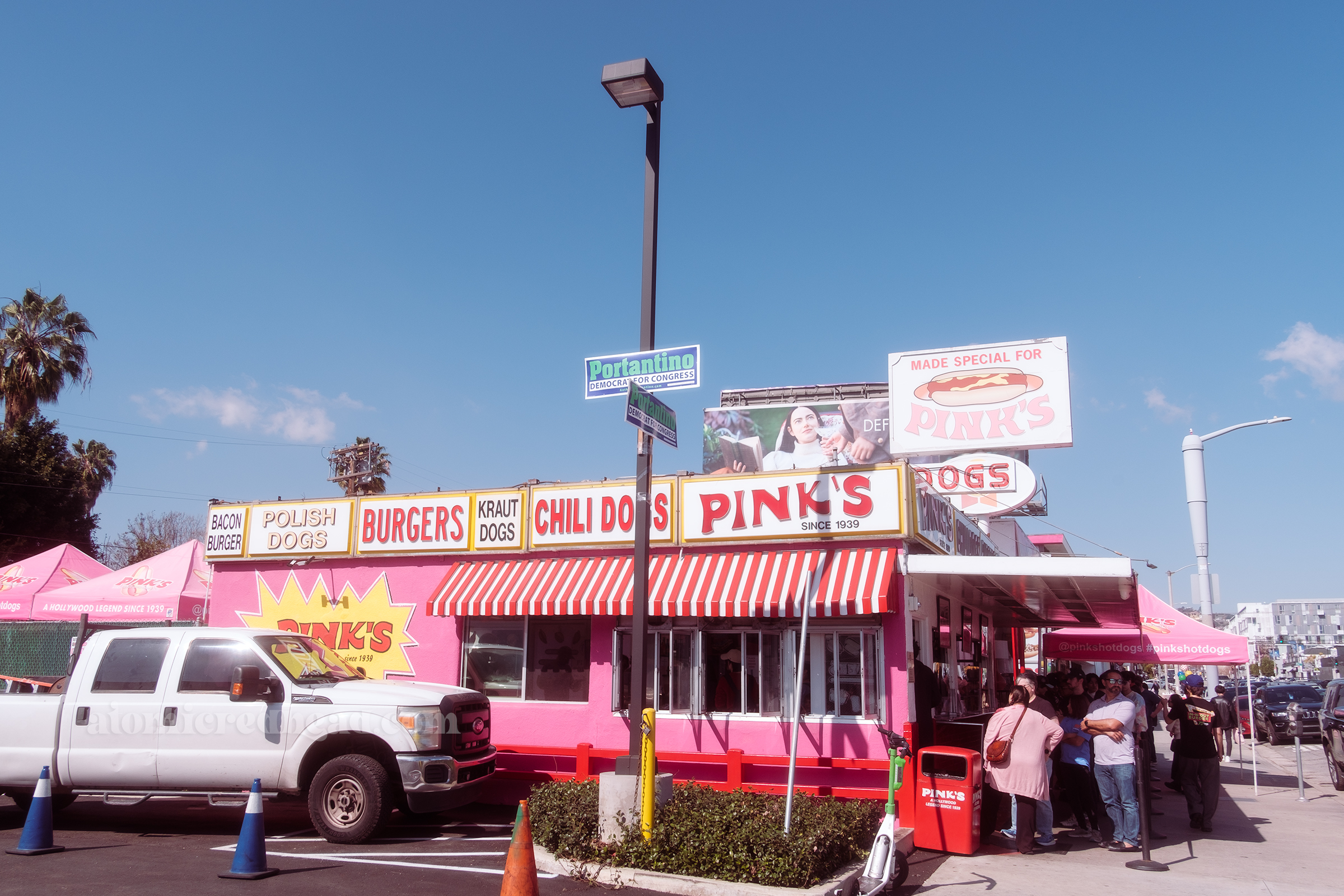 Overall view of Pink's. The large backlit plastic sign of a hot dog reads "Made Special for Pink's" with another backlit plastic sign below reading "Hoffy." Under an awning people line up to enter a small doorway to order. The side of the building feature a red and white stripe awning and lots of signs reading "Bacon Burger, Polish Dogs, Burgers, Kraut Dogs, Chili Dogs, Pink's Since 1939"