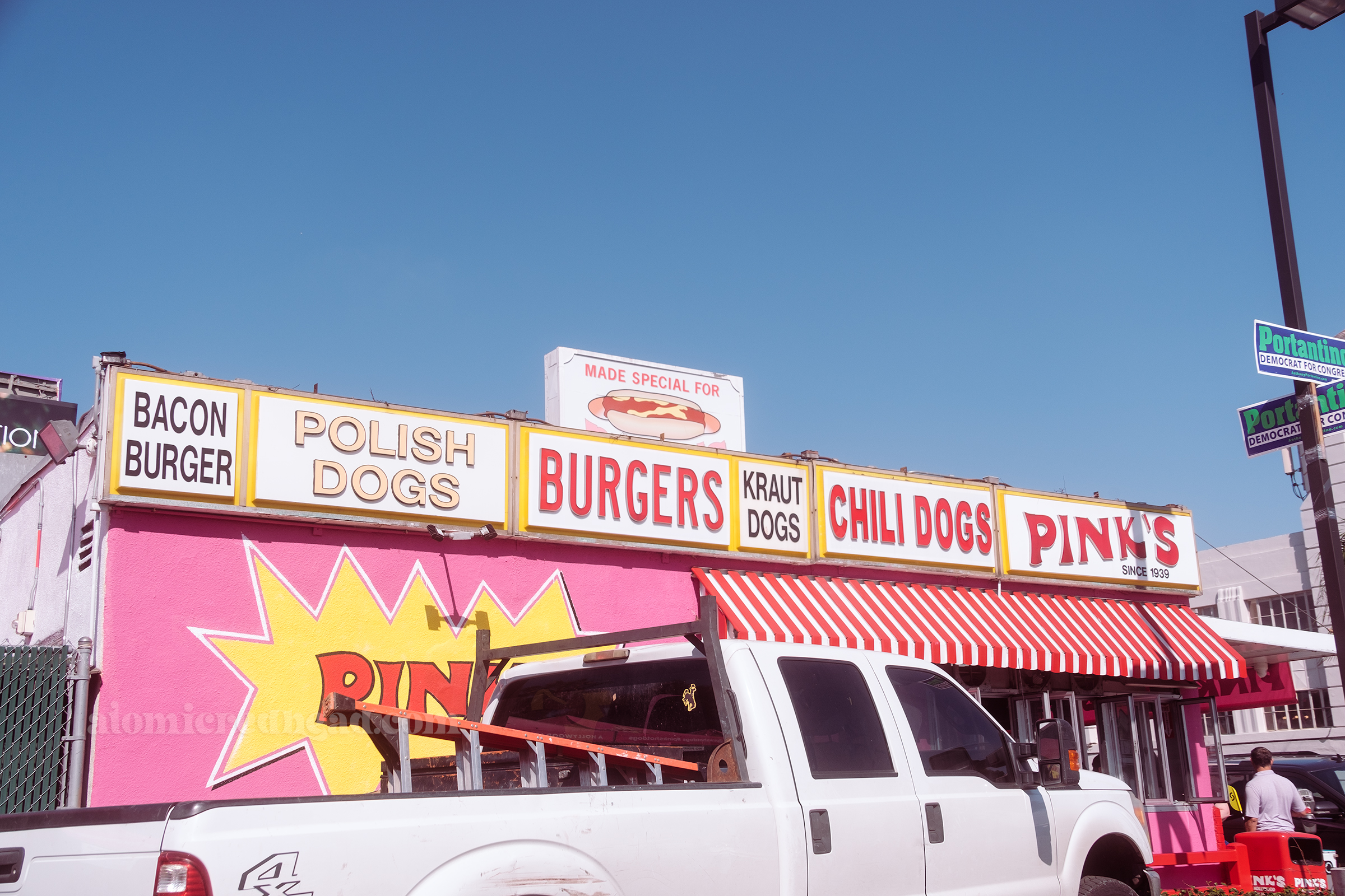 Side of the building is painted pink and features many signs reading "Bacon Burger, Polish Dogs, Burgers, Kraut Dogs, Chili Dogs, Pink's Since 1939"