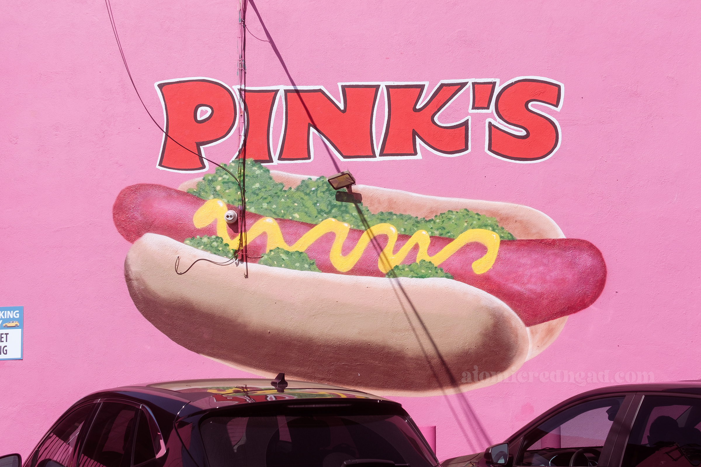 A wall is painted pink with a large mural of a hot dog with mustard and relish on it, above in bold red letters reads "Pink's"