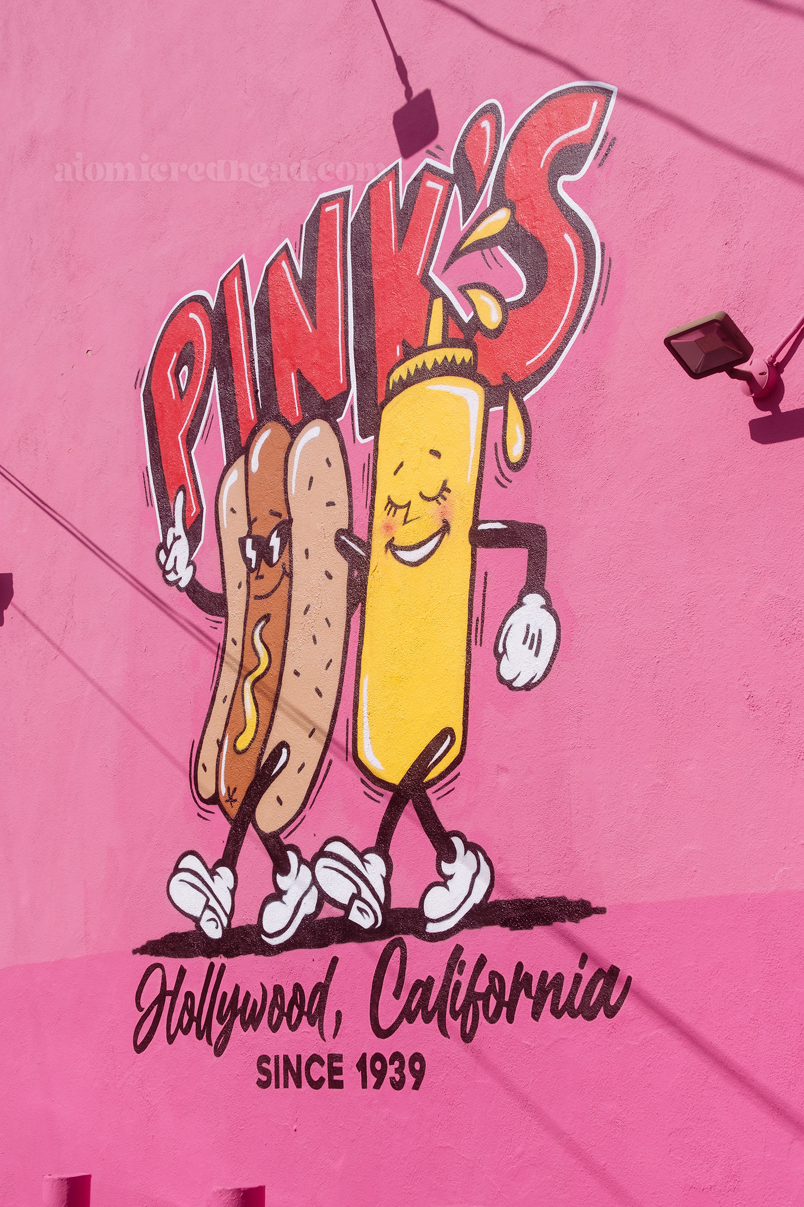 A mural of a hot dog and bottle of mustard walking together is painted on a pink wall, in letters across the top it reads "Pink's" and below reads "Hollywood, California Since 1939"