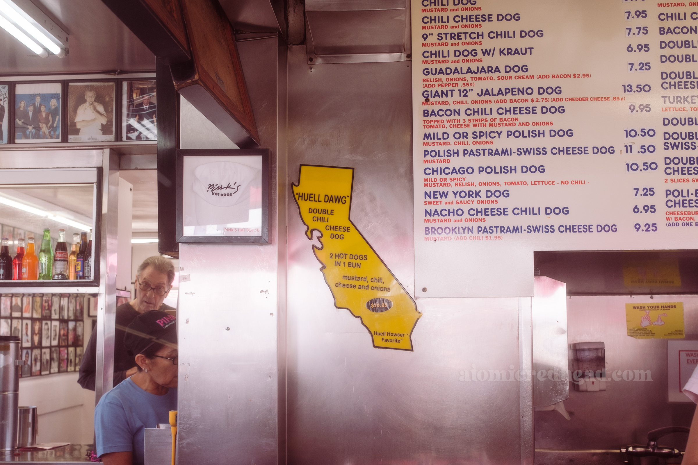A large yellow plastic sign in the shape of California reads "Huell Dawg Double Chili Cheese Dog 2 Hot Dogs in 1 Bun, Mustard, Chili, Cheese, and onions Huell Howser Favorite"