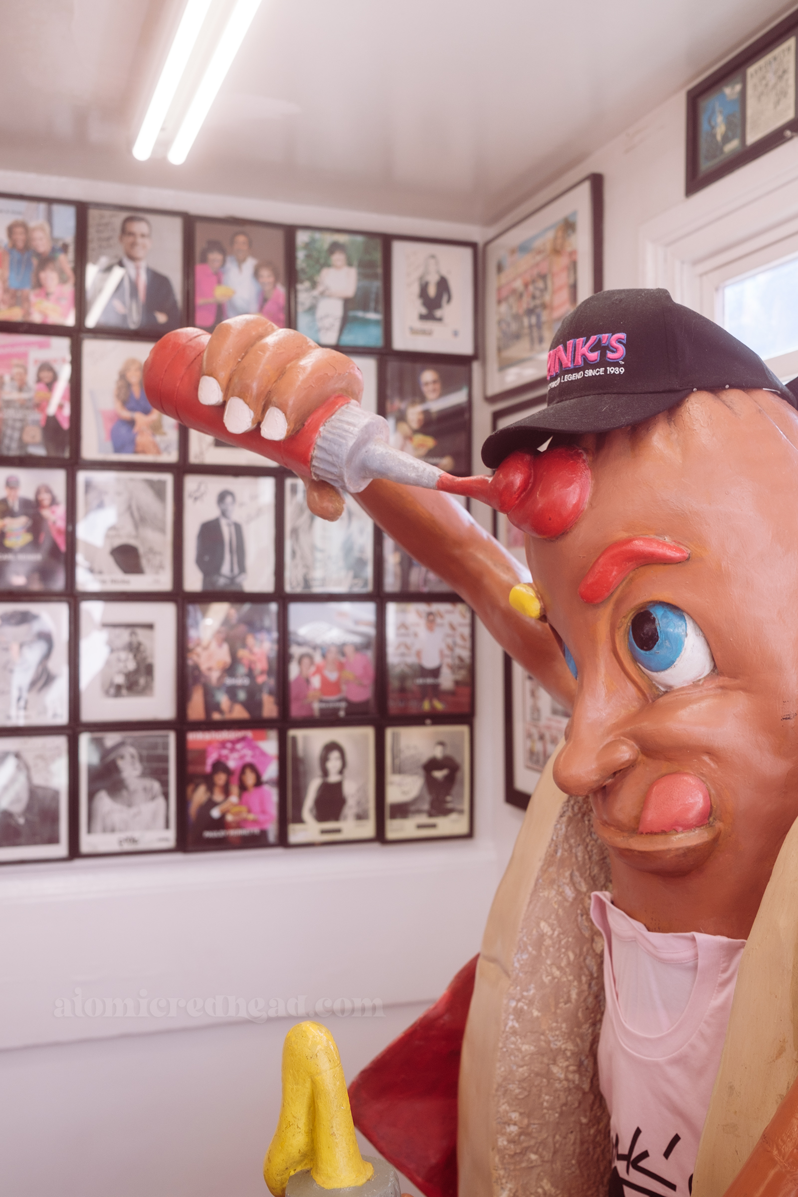 A large statue of a hot dog give himself some ketchup, behind him the wall is lined with photos of celebrities.