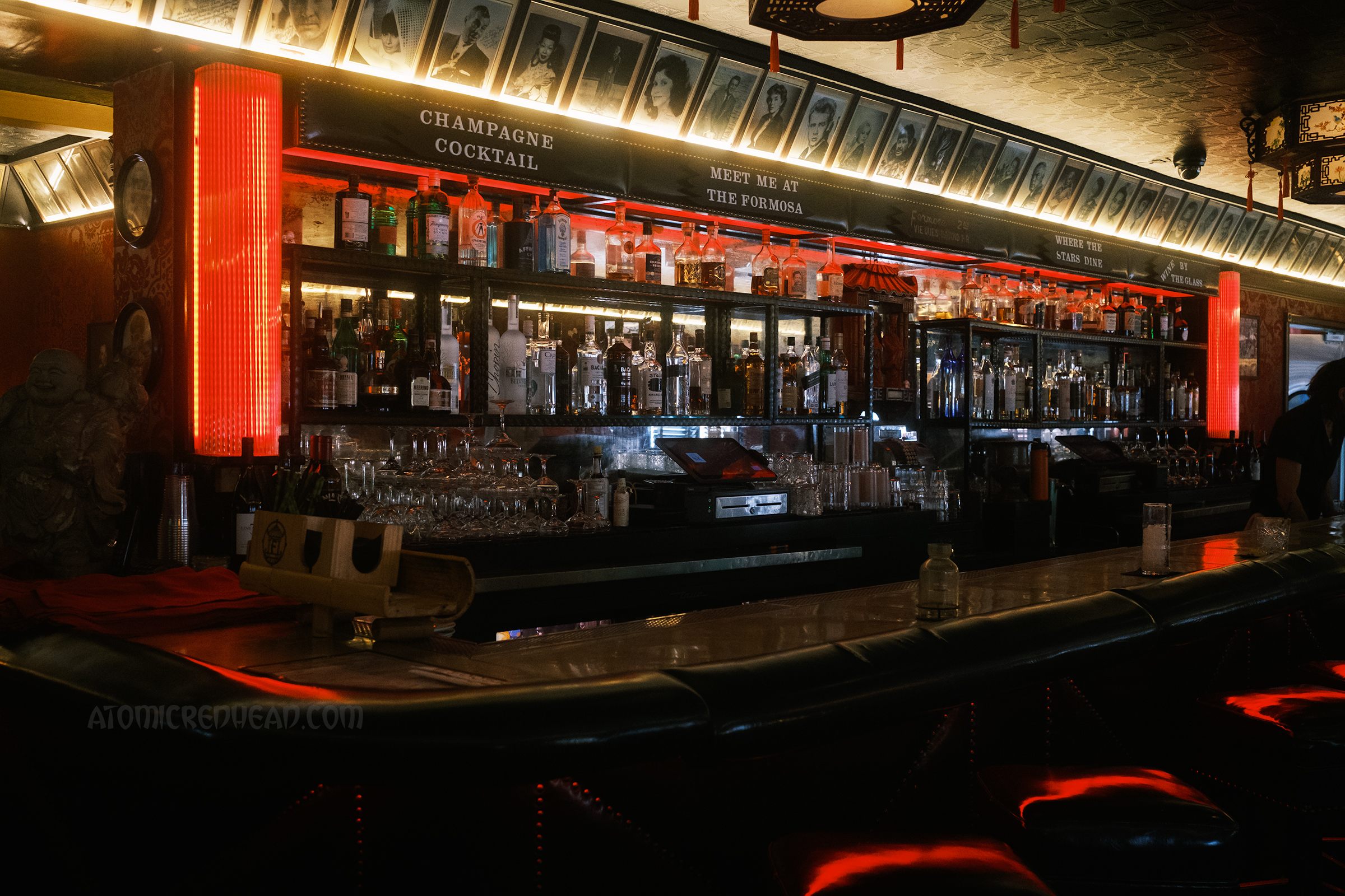Overall view of the bar, black and white photos of stars hang at the top, with small signs reading "Champaign Cocktail" and "Meet Me at the Formosa" the bar features red lights and bottles of booze.