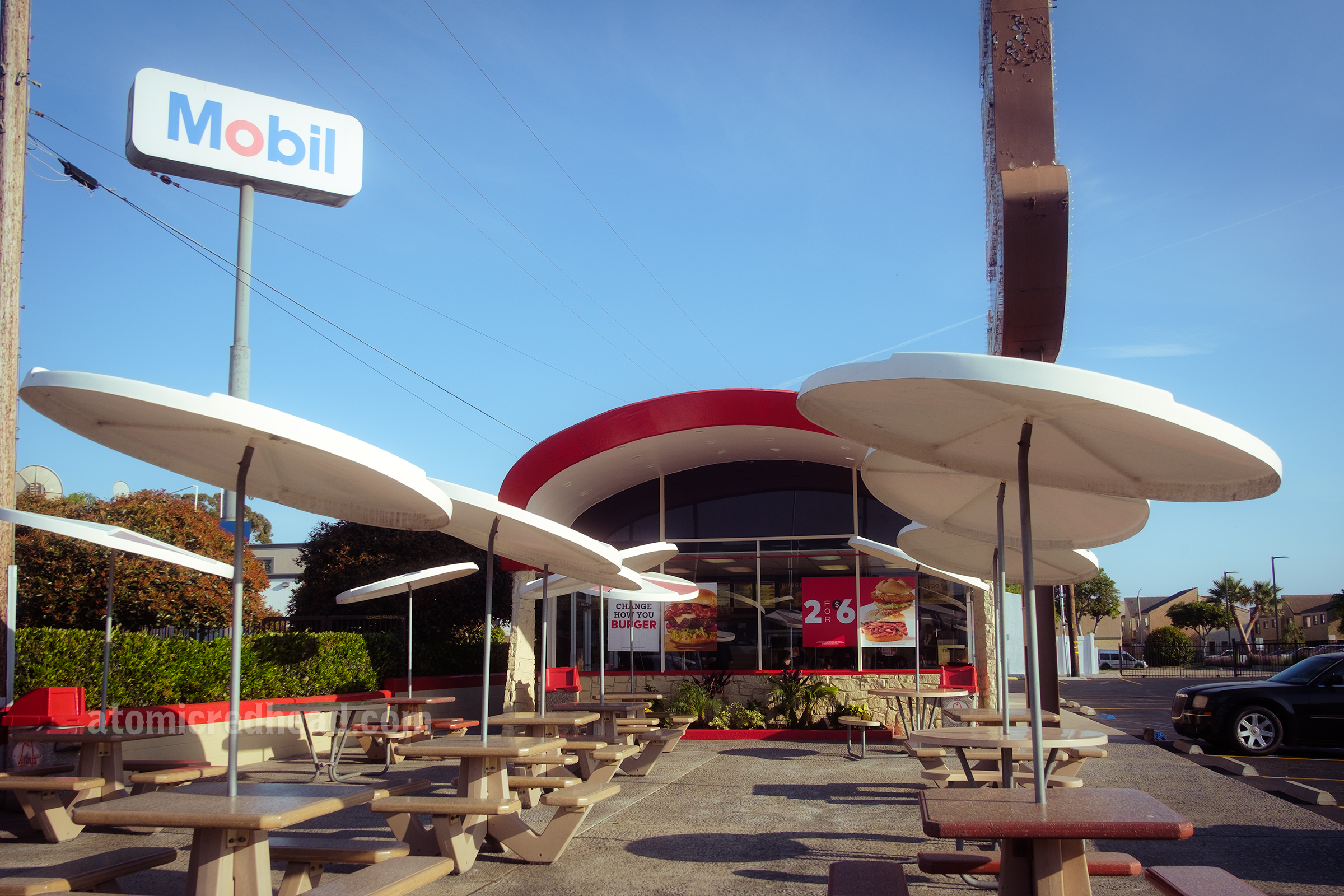 Front of the Arby's, with a curved red roof, and almost floor to ceiling wall of glass. A collection of small outdoor tables feature circular sold umbrellas of white.