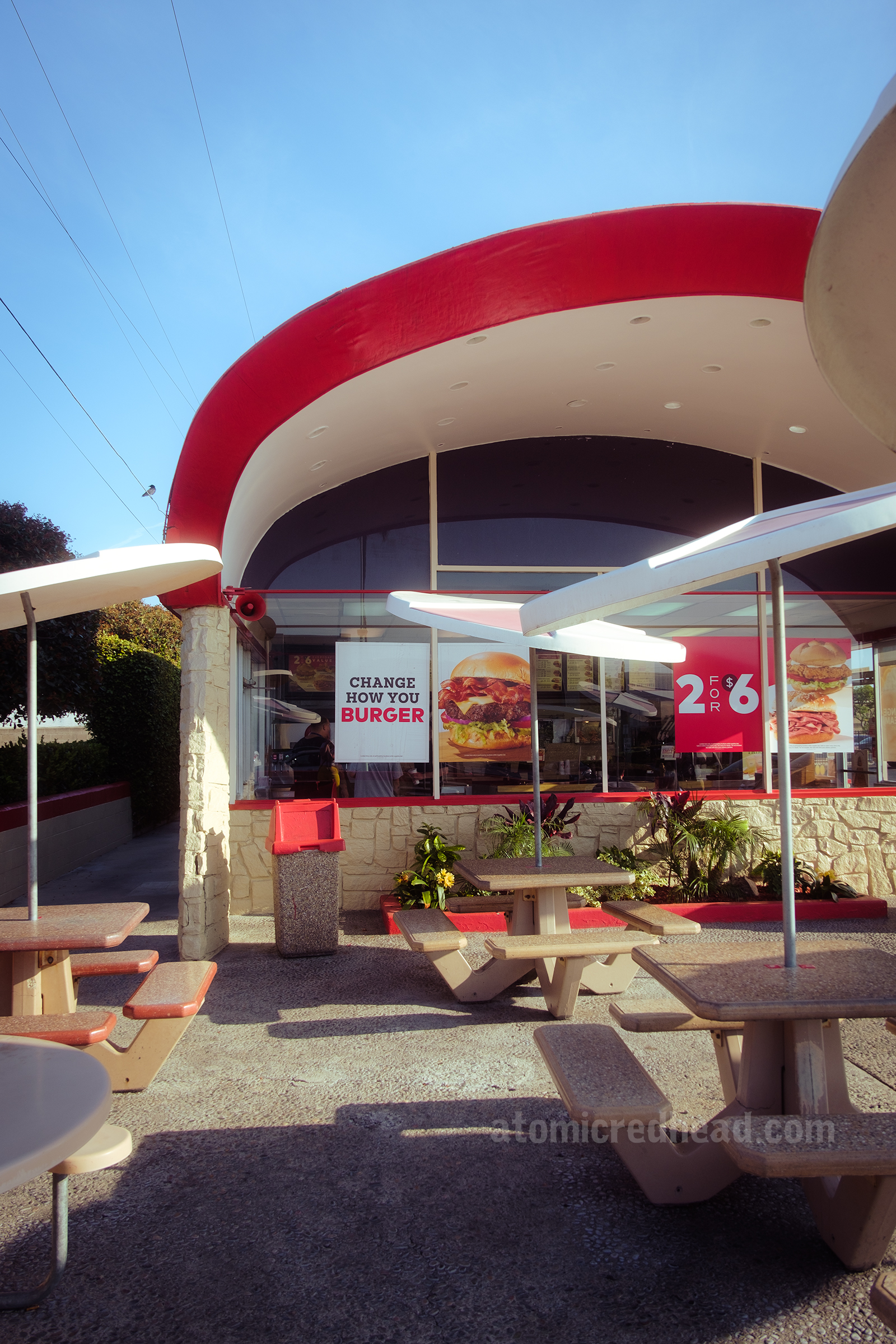 Front of the Arby's, with a curved red roof, and almost floor to ceiling wall of glass. A collection of small outdoor tables feature circular sold umbrellas of white. The front of the building juts out a few feet, creating an awning. The underside is painted white and has recessed lighting.