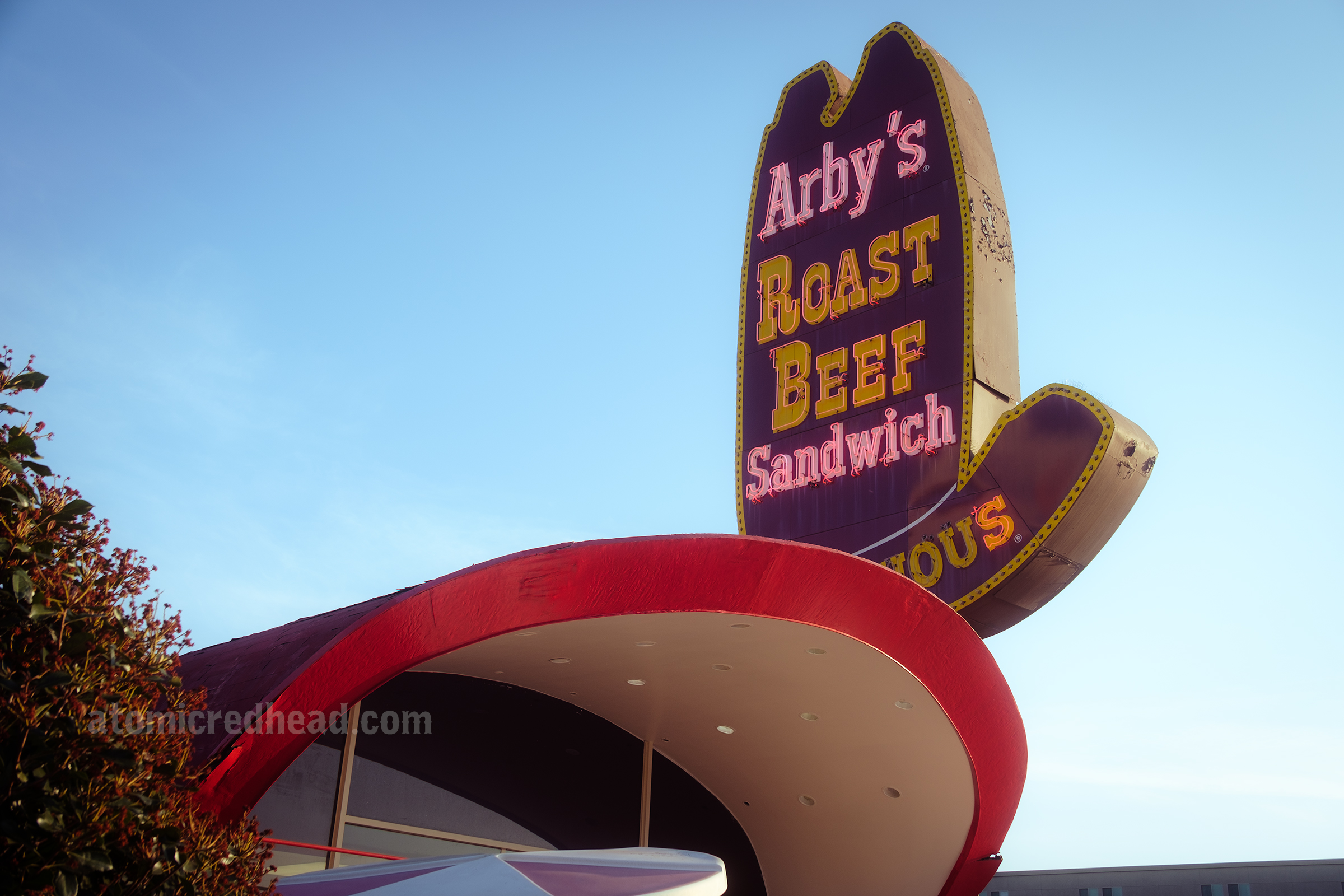 Angled view of the front of the building with its curved red roof. Behind it is the massive ten gallon hat sign that has yellow and white letters reading "Arby's Roast Beef Sandwich"
