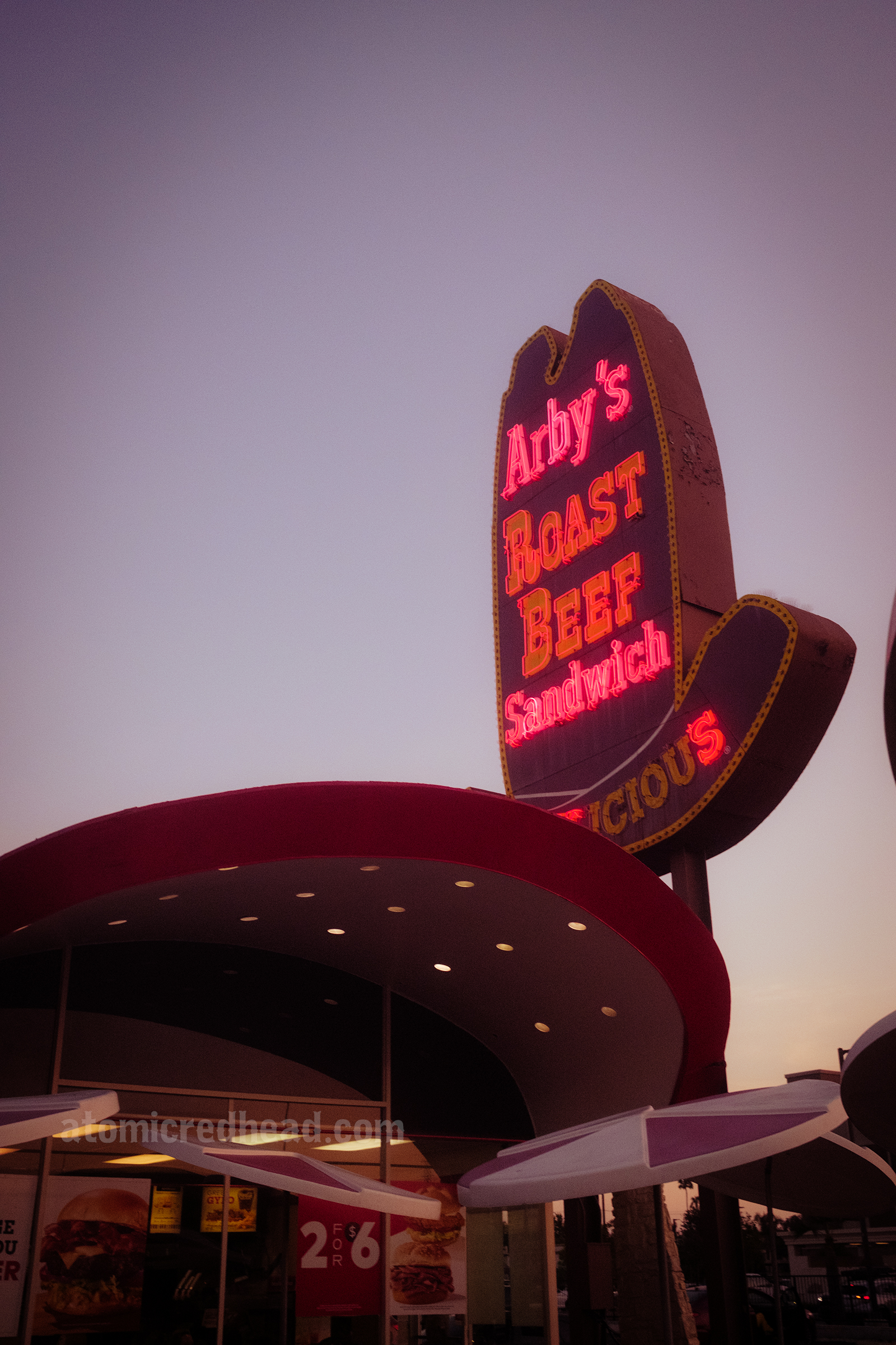 Angled view of the front of the building with its curved red roof. Behind it is the massive ten gallon hat sign that has yellow and white letters reading "Arby's Roast Beef Sandwich" lit up at night in neon.