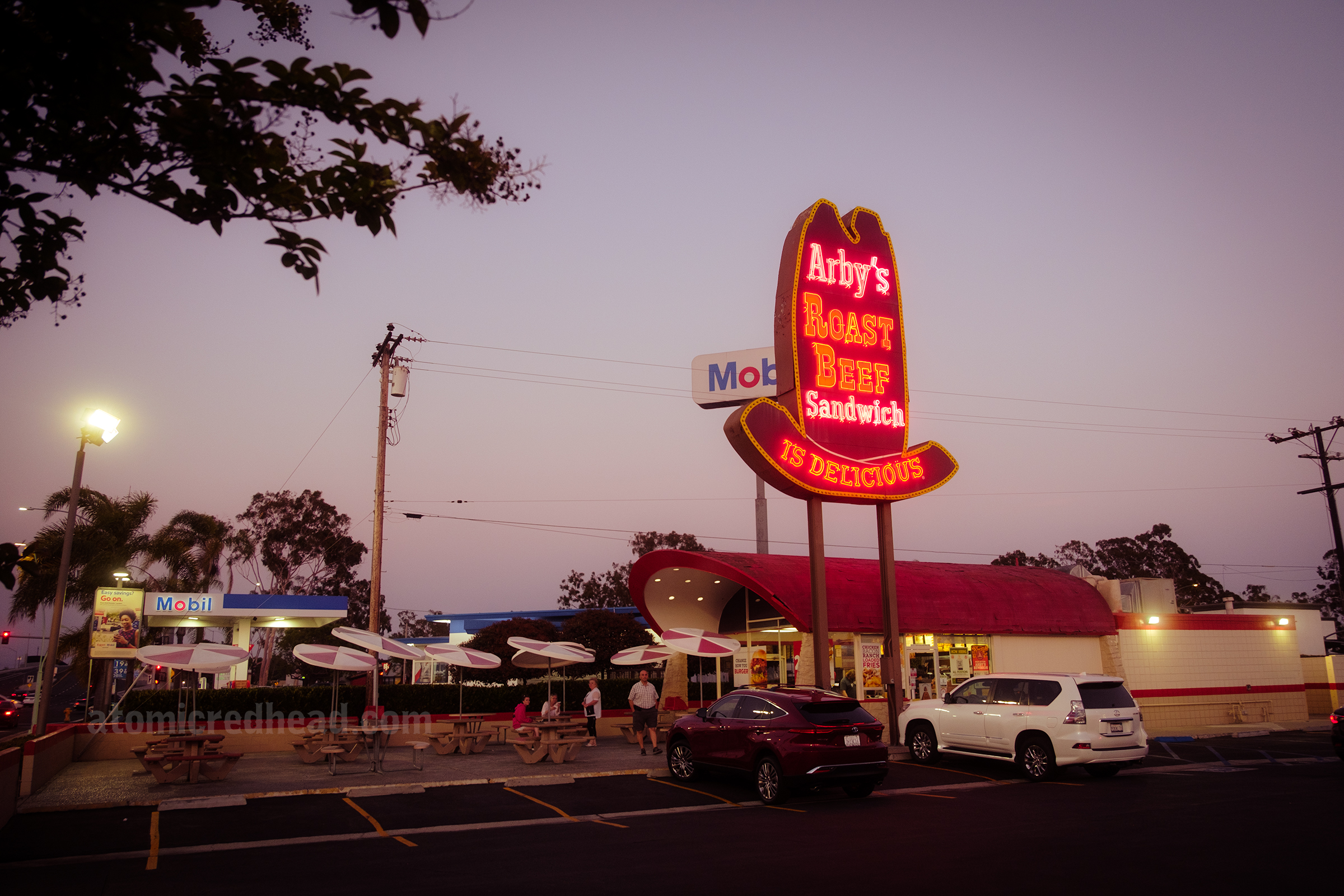 Overall view of the restaurant, with the towering sign, covered wagon style building and patio seating, at dusk, with a purple-pink sky and the sign lit up with neon.
