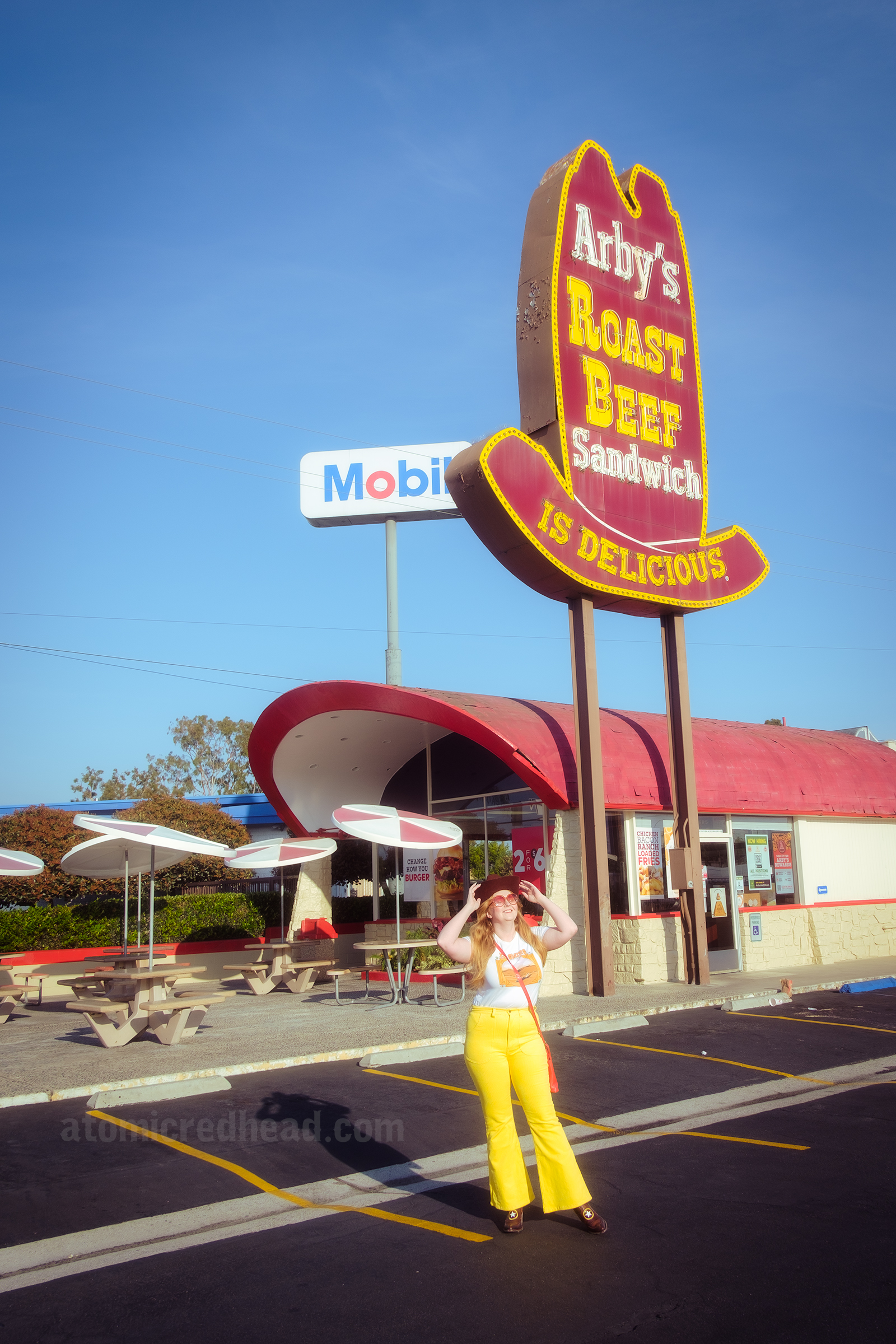 Overall view of the restaurant, with the towering sign, covered wagon style building and patio seating. In the parking lot I stand in front of the sign, wearing a brown cowboy hat, white shirt, and yellow pants.