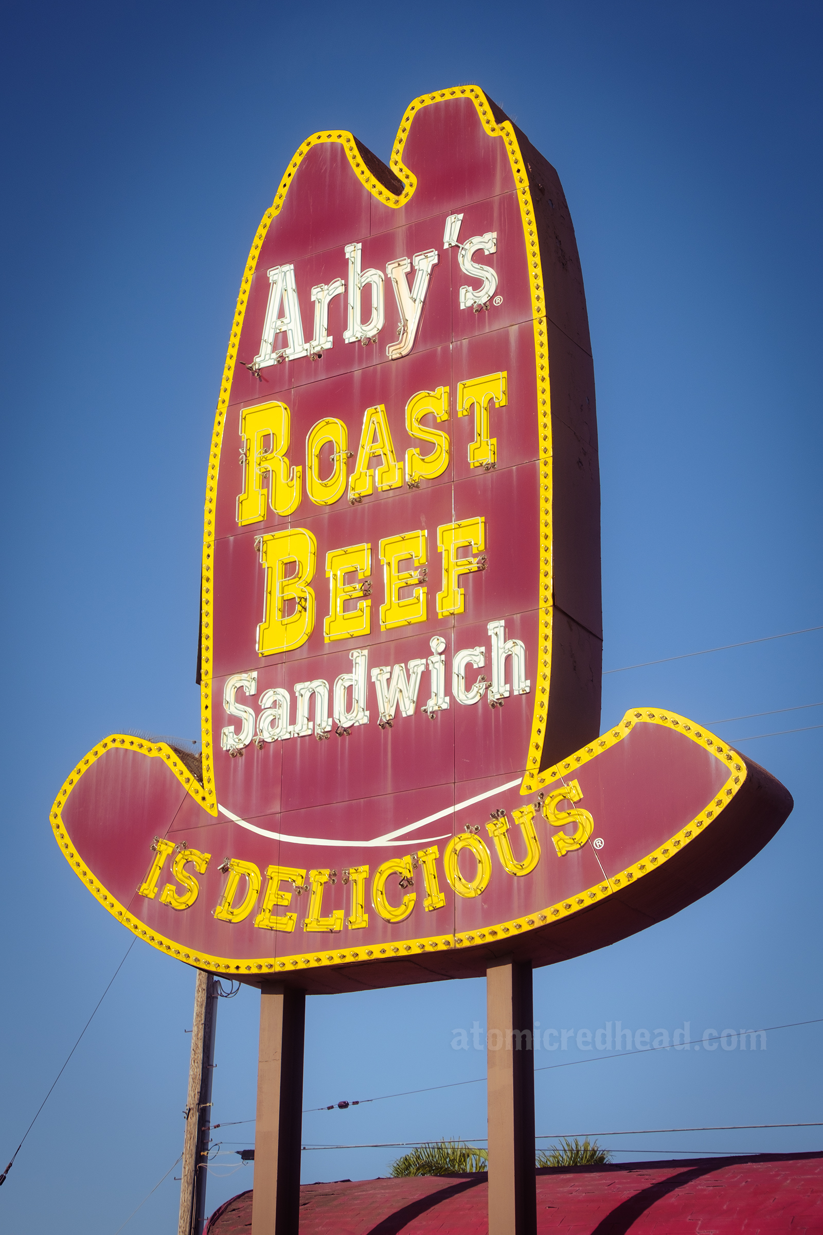 Close-up view of the entire neon sign made to look like a tall ten gallon style cowboy hat. "Arby's" is in white painted letters, "Roast Beef" in yellow painted letters, "Sandwich" in white letters and "is delicious" in yellow letters.