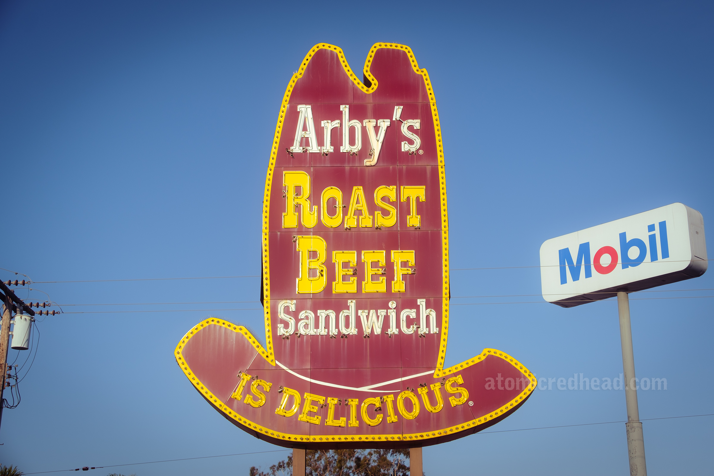 Close-up view of the entire neon sign made to look like a tall ten gallon style cowboy hat. "Arby's" is in white painted letters, "Roast Beef" in yellow painted letters, "Sandwich" in white letters and "is delicious" in yellow letters.