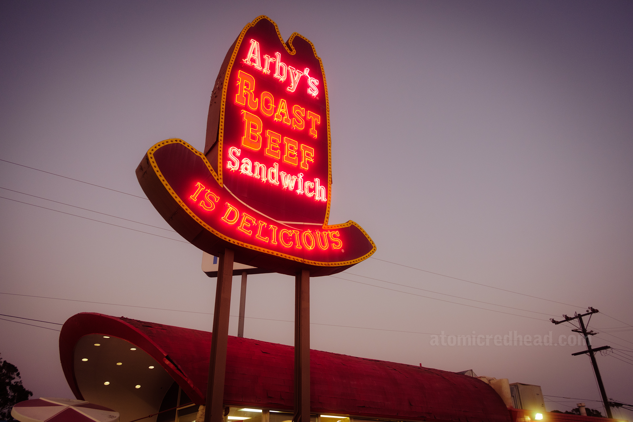 Angled view of the ten gallon hat sign that is lit up with neon reading "Arby's Roast Beef Sandwich is Delicious" below is the curved roof of the restaurant.