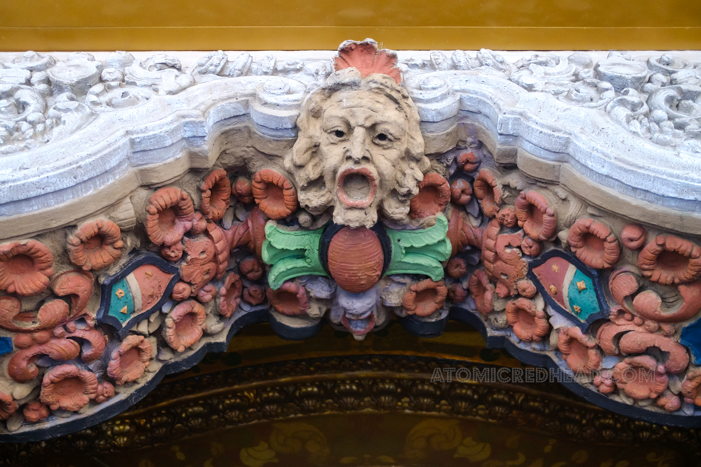 Sculpted detail above the box office, of a face with an open mouth. To the left and right are red flowers.