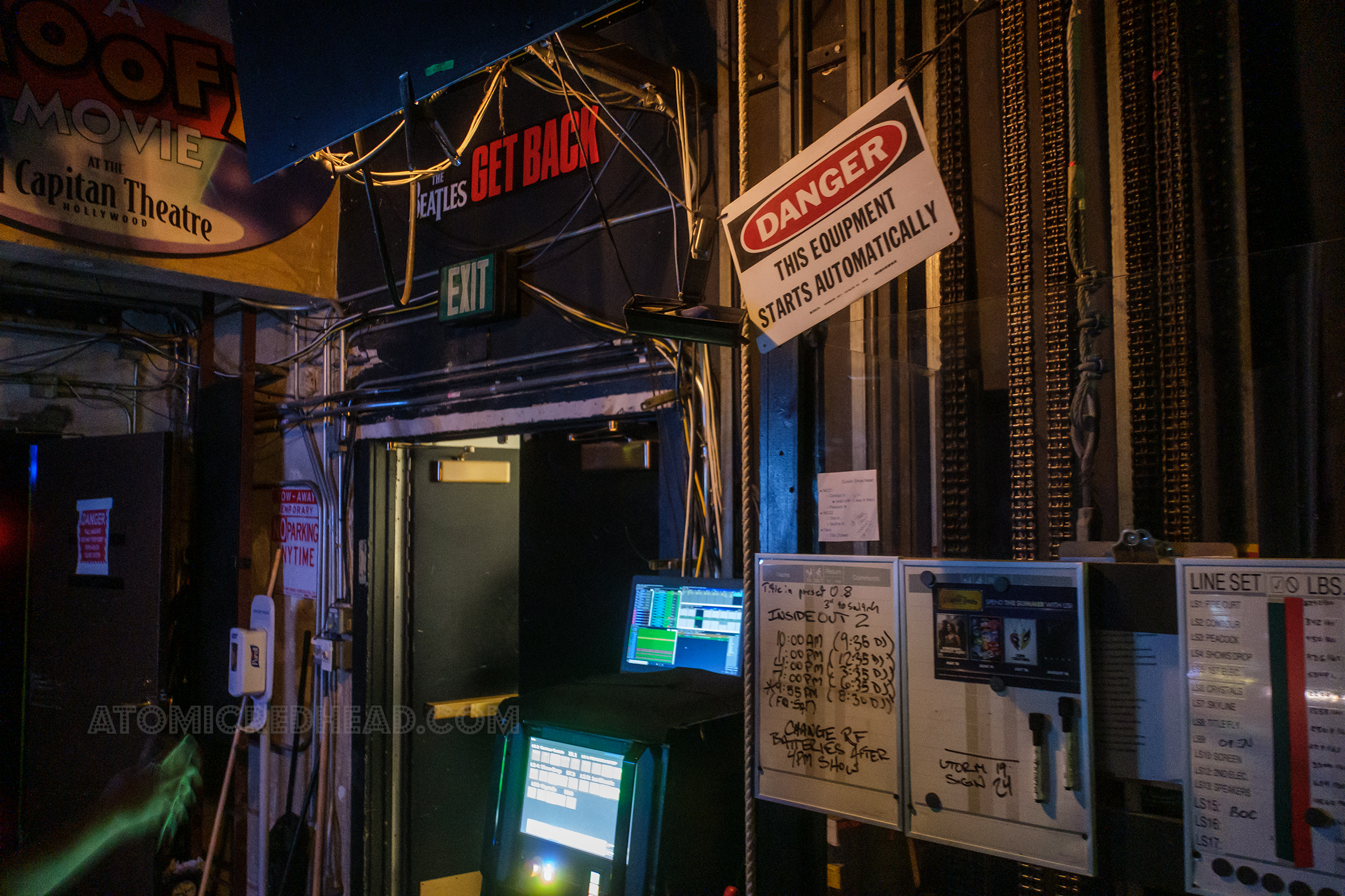 A computer sits near a doorway and by the fly rail. Hanging above is a black, white, and red sign that reads "Danger this equipment starts automatically"